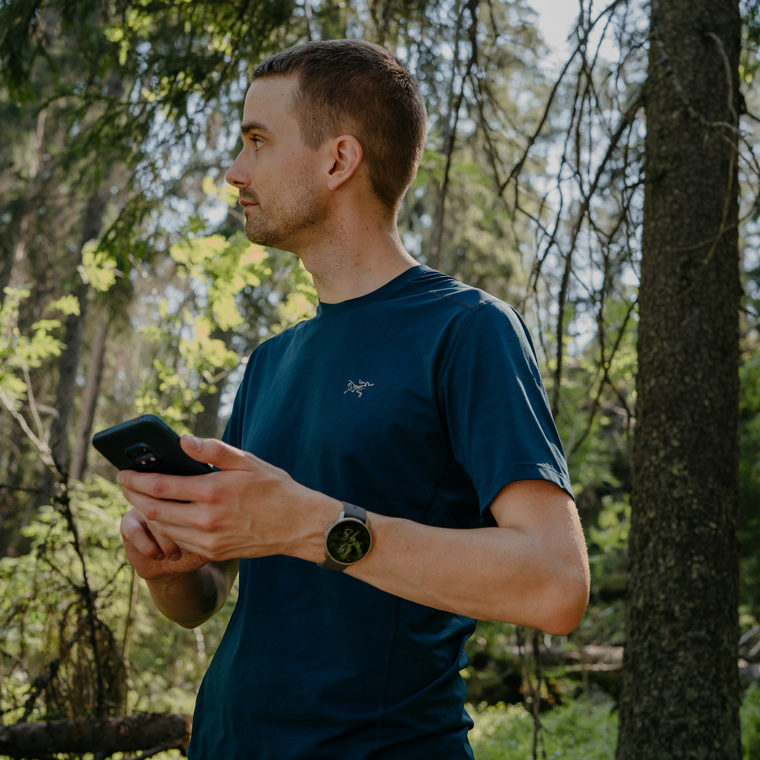 Hombre con camisa azul sosteniendo un teléfono en un bosque, con un reloj en la muñeca.