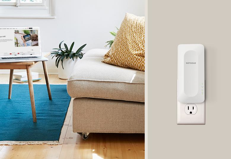 Un répéteur WiFi Netgear branché sur une prise murale, avec un ordinateur portable et un canapé en arrière-plan.