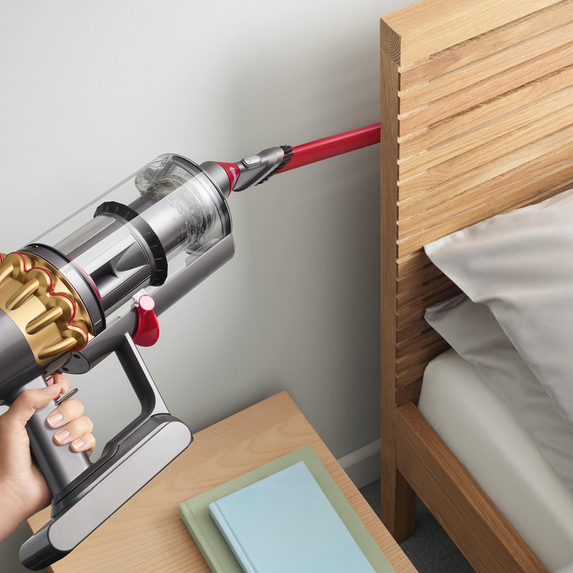 Eine Person saugt mit einem Dyson-Staubsauger in der Nähe eines Bettes. Der Staubsauger ist silber und rot.