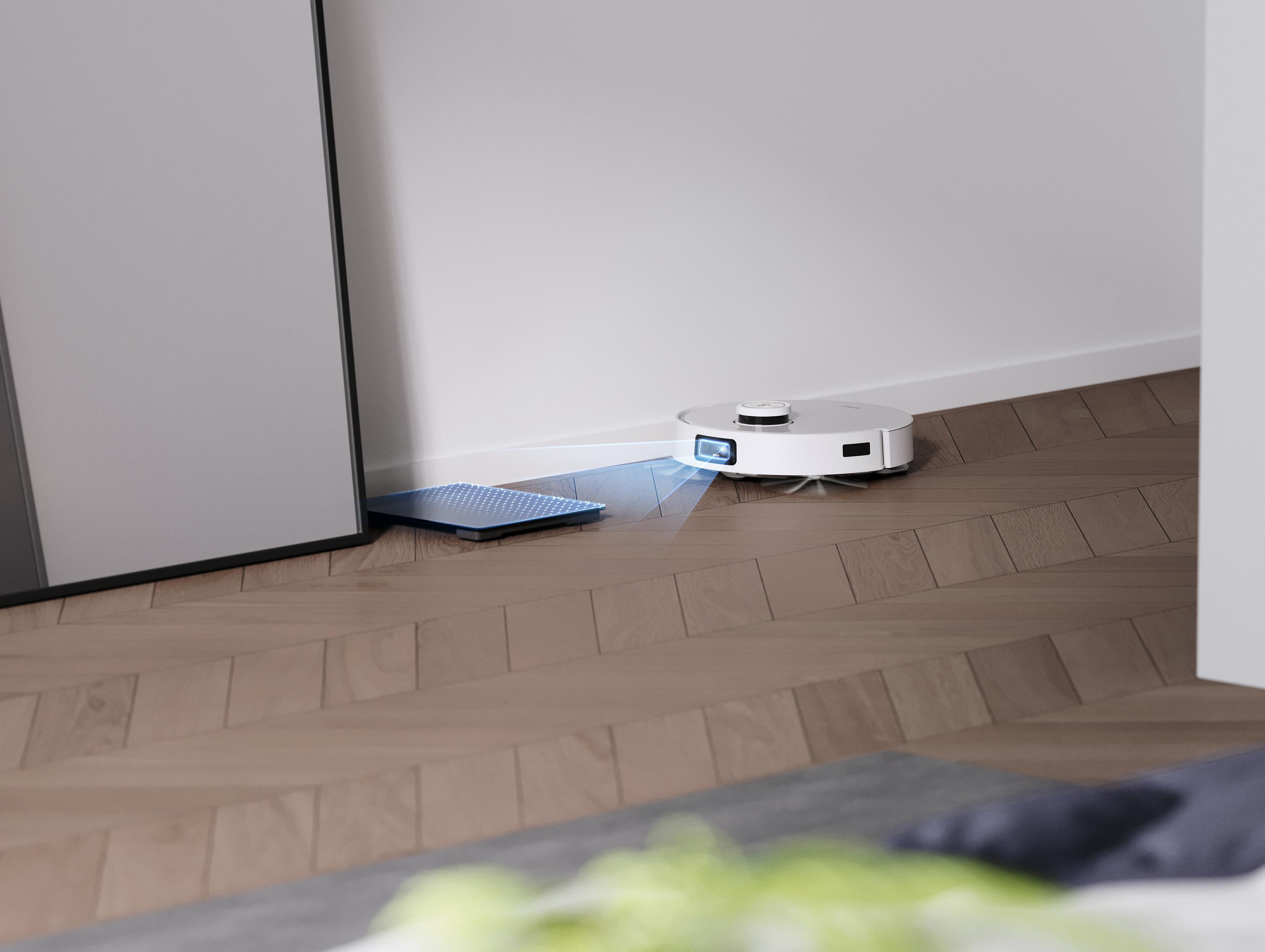 Weißer Roboter-Staubsauger reinigt einen Holzboden neben einer Wand.