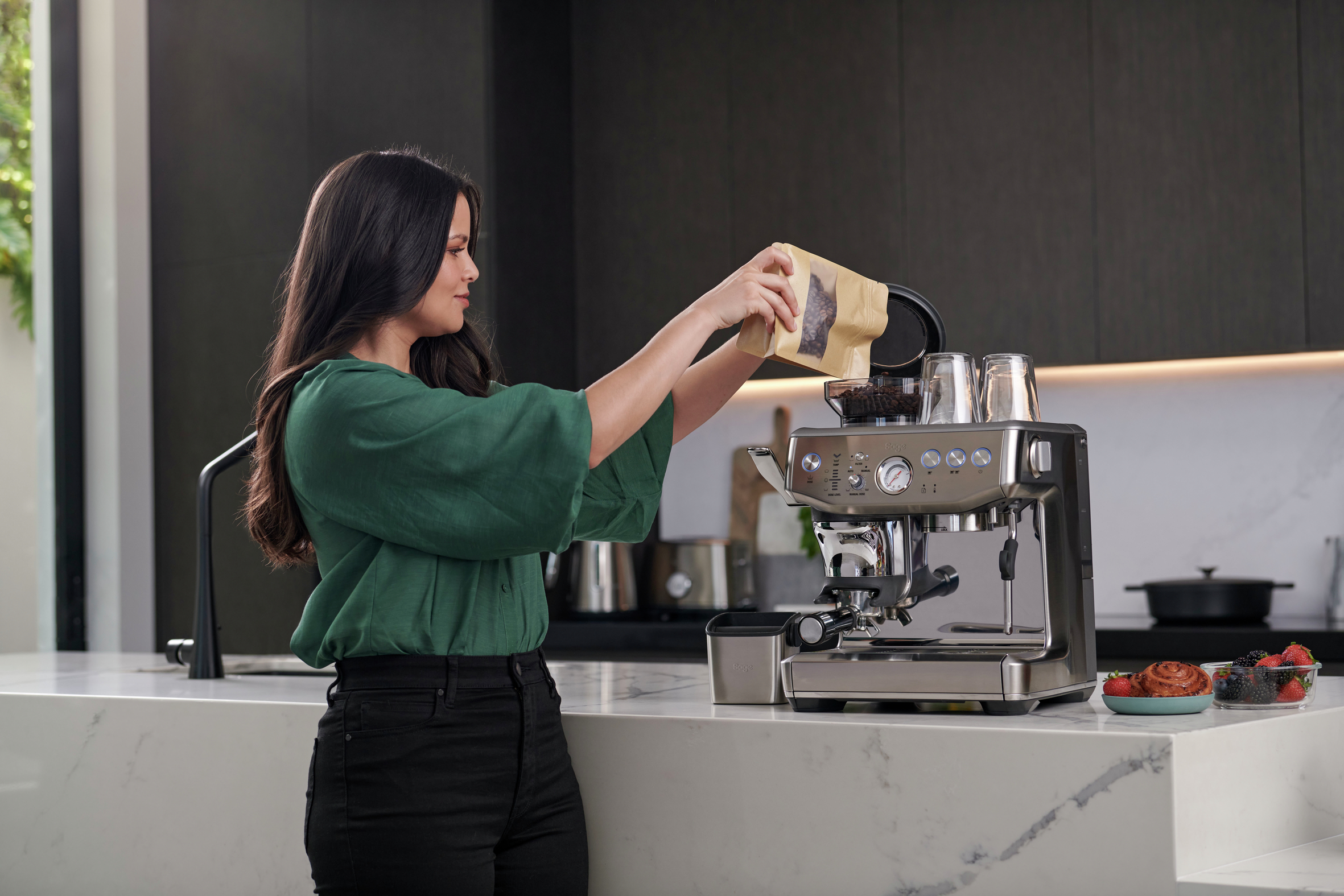 Frau gießt Kaffeebohnen in eine silberne Espressomaschine auf einer Arbeitsplatte. Küchenszene.