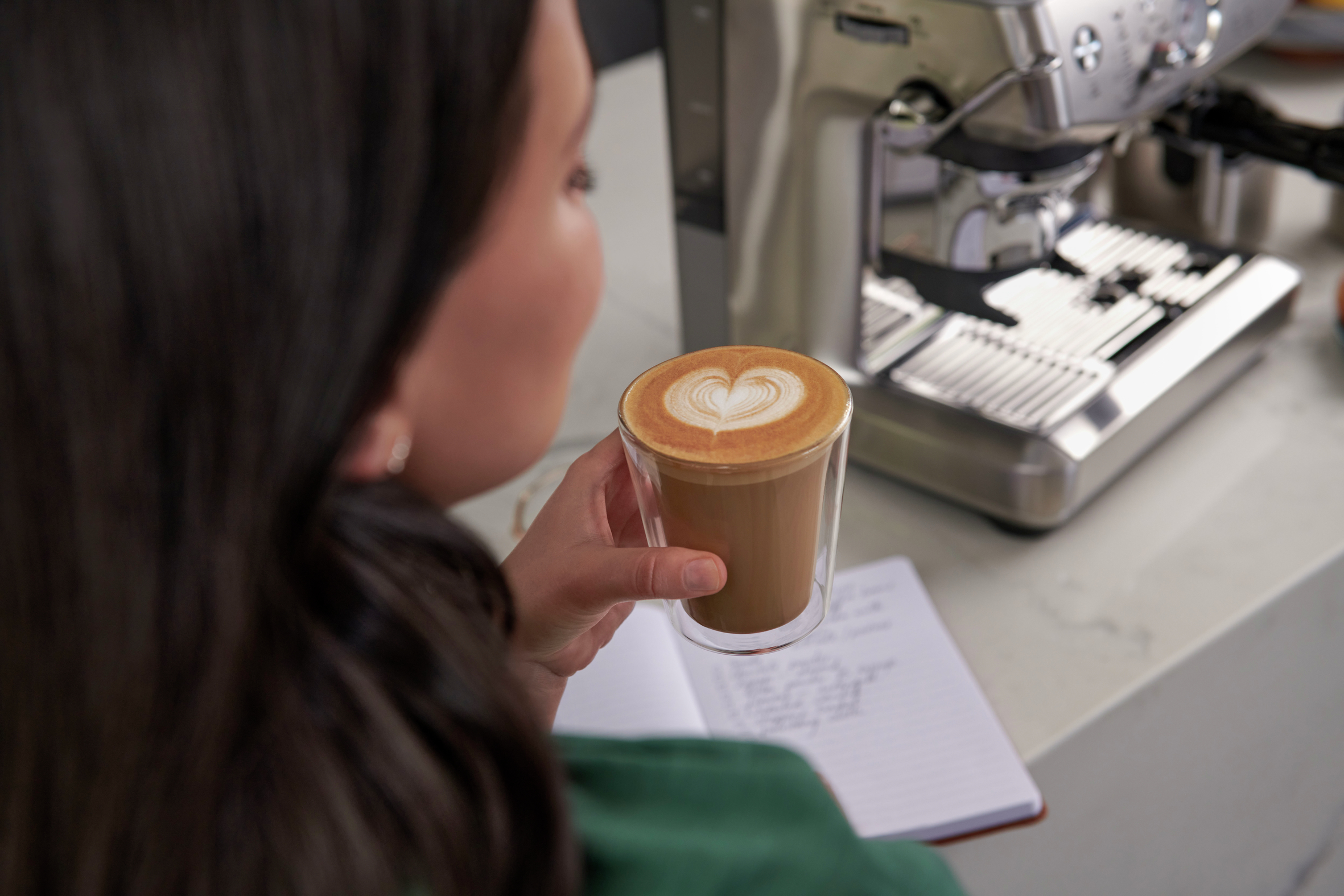Frau hält Latte mit Herzmuster, neben Notizbuch und Kaffeemaschine.
