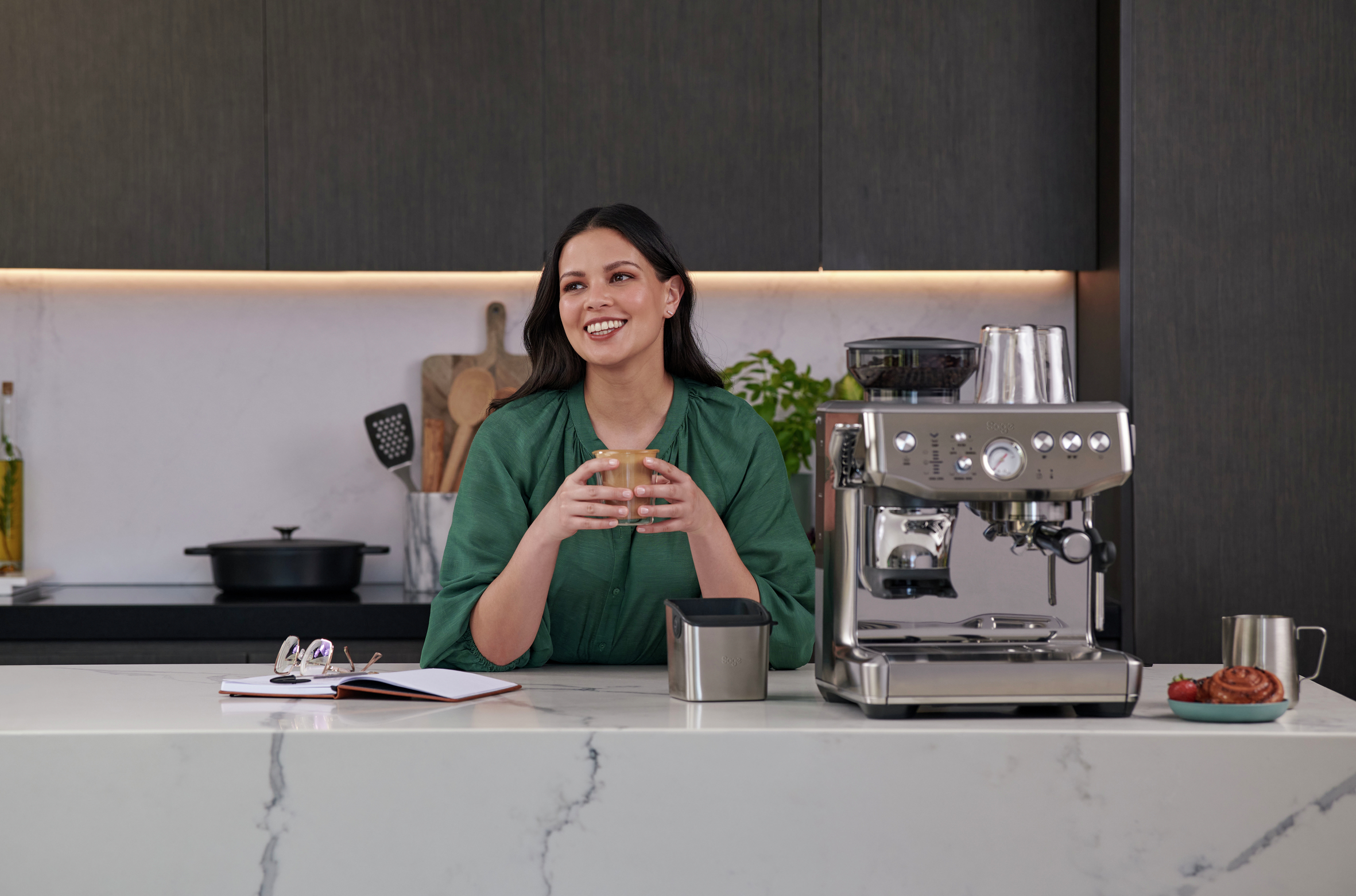 Frau lächelt, hält ein Getränk in einer Küche mit Kaffeemaschine.