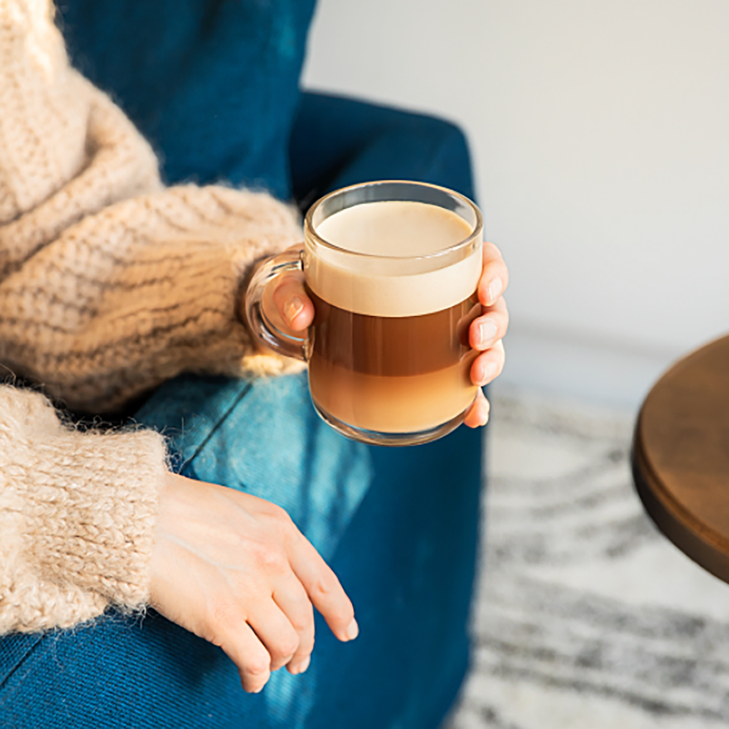 A person holding a glass mug of layered coffee with foam, seated in a blue chair.