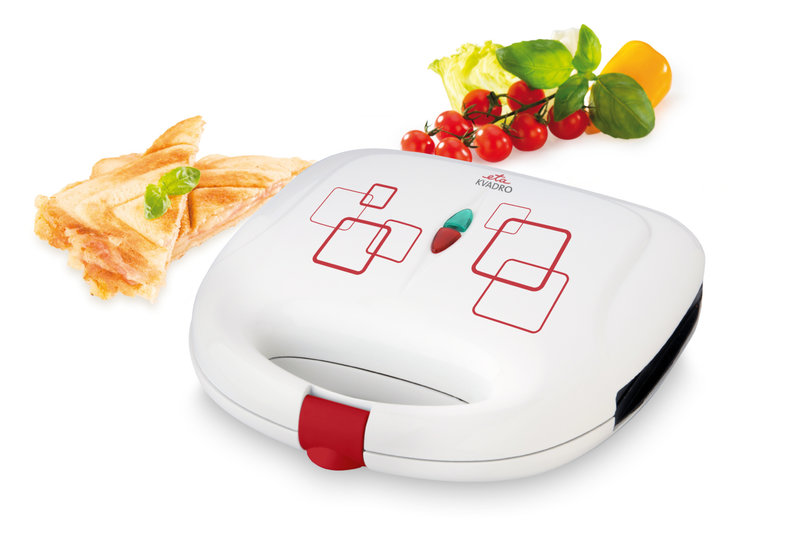 Egy fehér szendvicssütő és egy pirított szendvics látható a képen. A háttérben paradicsom, bazsalikom és egy sárga kaliforniai paprika látható.