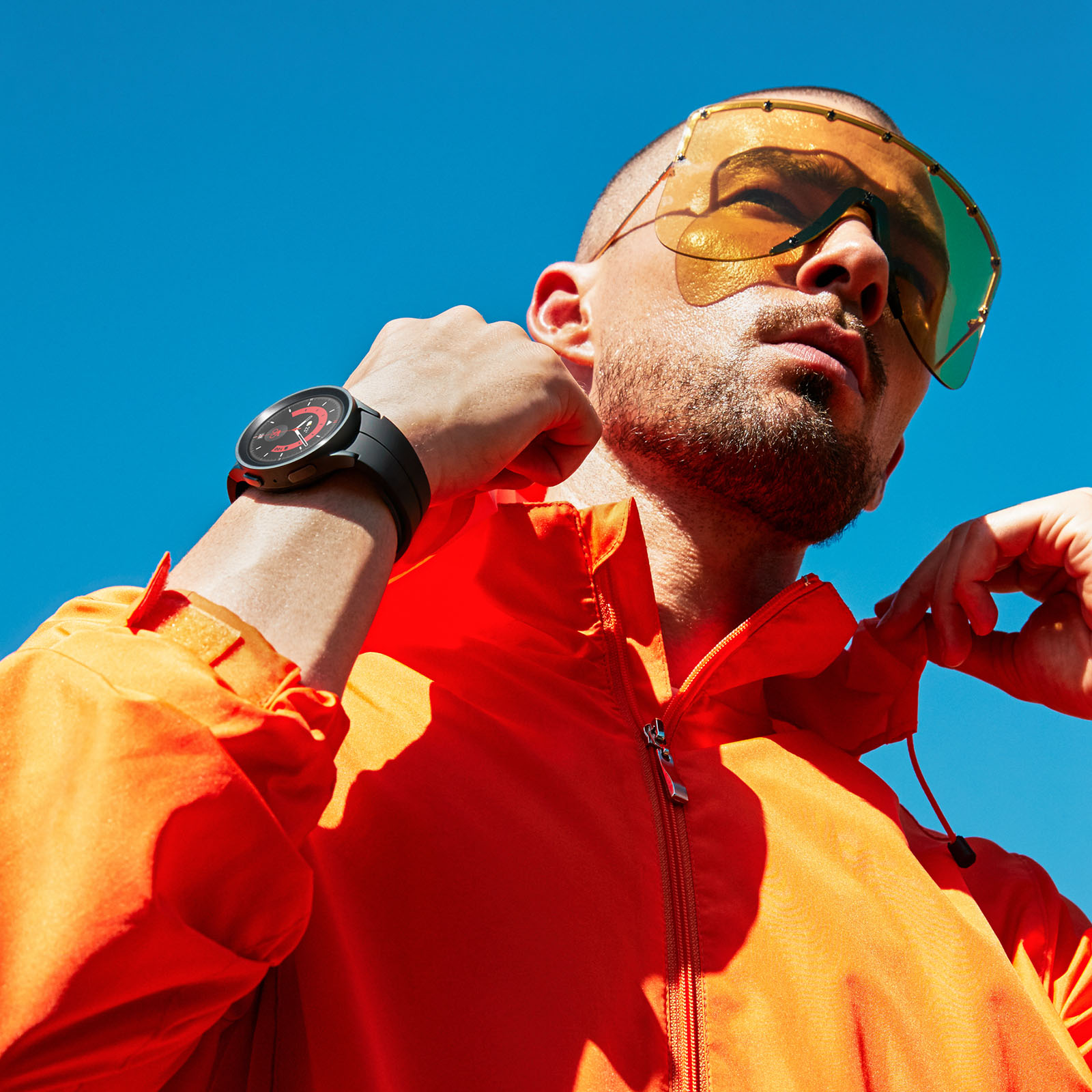 Mann in orangefarbener Jacke und Sonnenbrille, blickt in den blauen Himmel. Schwarze Uhr.