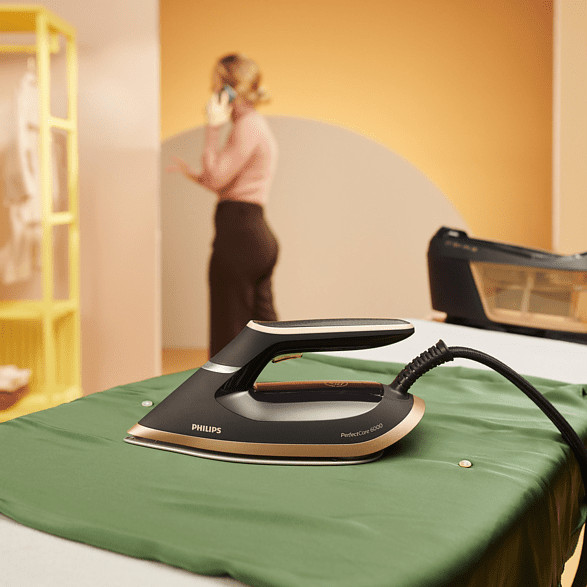 Table à repasser avec un tissu vert. Un fer à repasser noir et doré est sur le tissu. Femme au téléphone en arrière-plan.