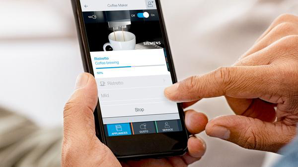 Person steuert Kaffeemaschine mit Telefon. Telefon zeigt Kaffeezubereitungsoptionen.