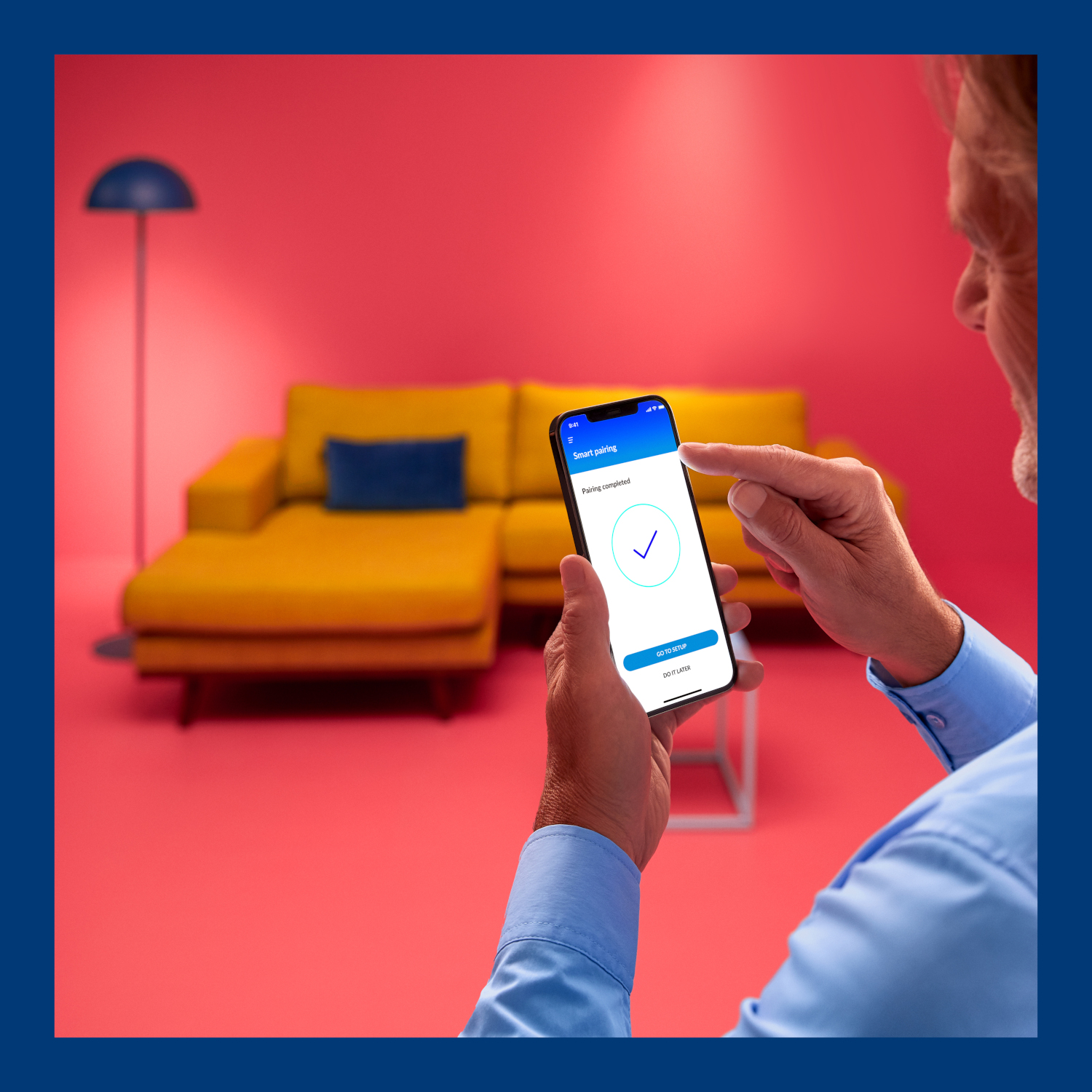 Een persoon houdt een telefoon vast die een gekoppeld apparaat weergeeft. De achtergrond is een gele bank en een roze muur.