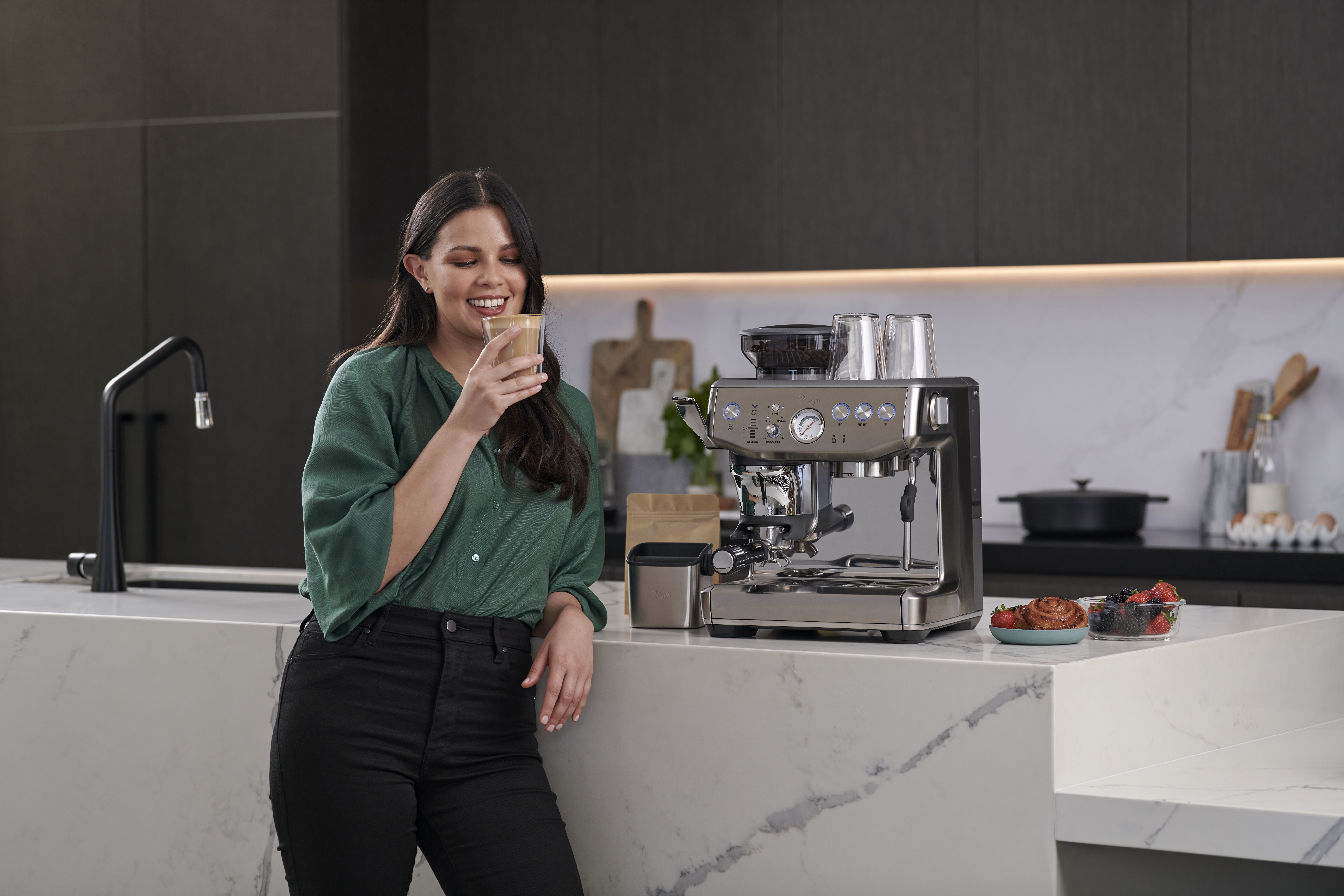 Femme avec machine à café, souriante, tenant un verre de café.