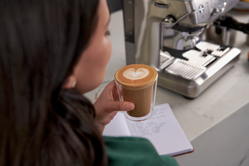 Femme tenant du café avec du latte art, à côté d'un cahier et d'une machine à café.