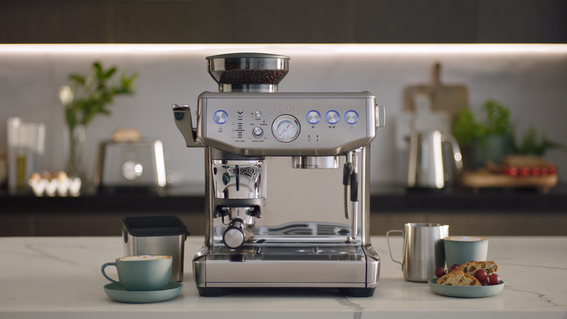 Une machine à café argentée sur un comptoir de cuisine. Il y a des grains de café et une tasse à côté.