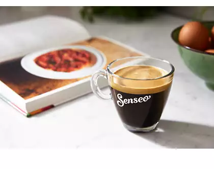 Een glazen kopje koffie met het woord 'Senseo', geplaatst bij een open kookboek.