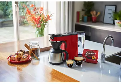 Een keukenscène met een rode koffiezetter, koffie en gebak op een wit aanrecht.