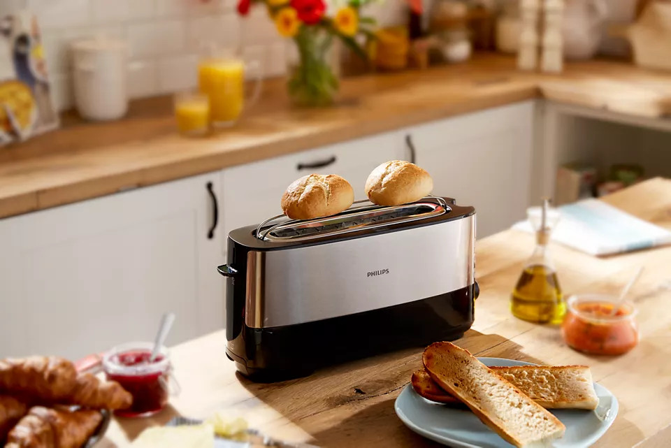 Toaster with two toasted rolls. Bread and jam are on the table, wooden counter in the background.