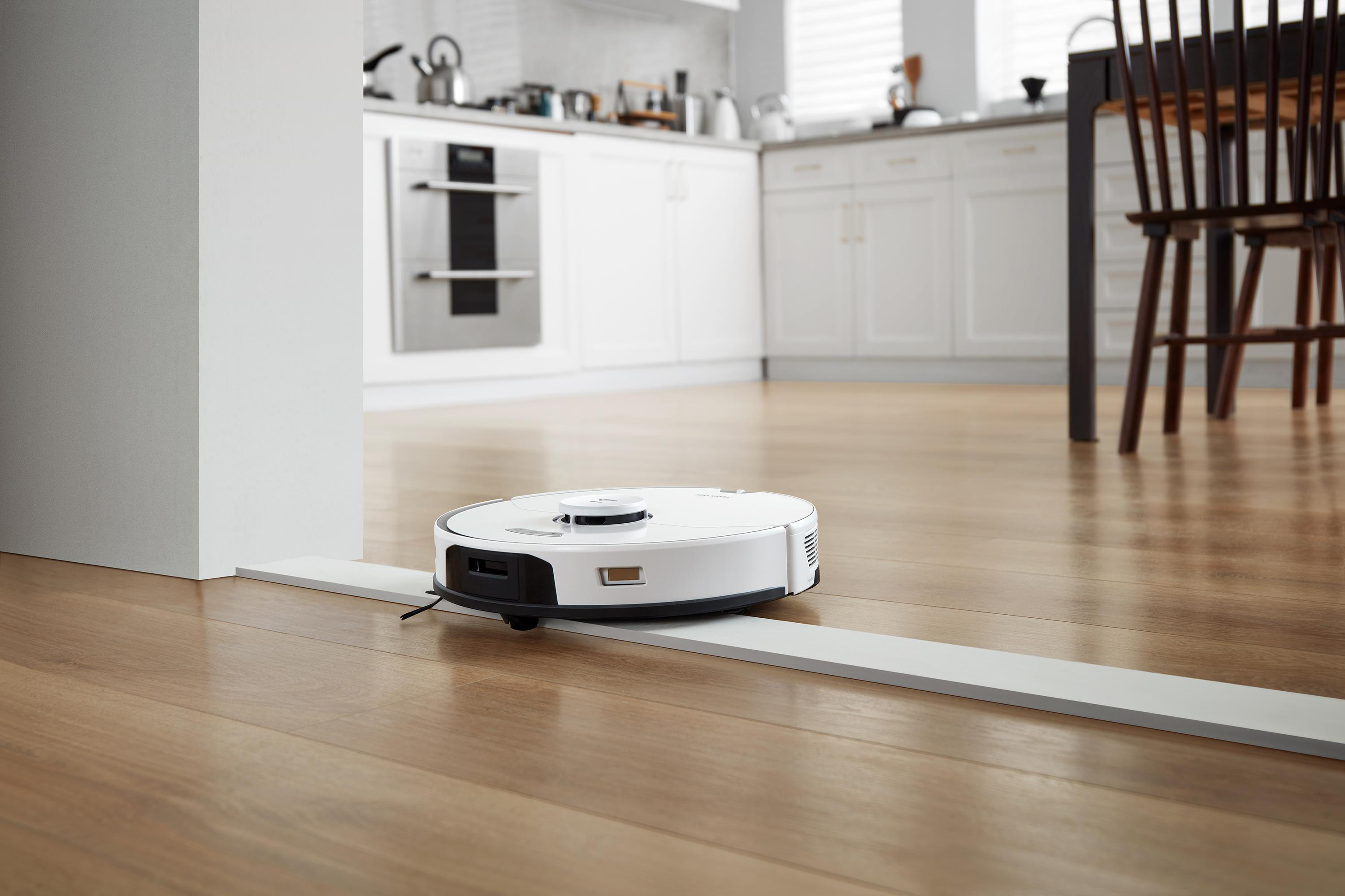 Weißer Roboter-Staubsauger auf einem Holzboden in einer Küche.