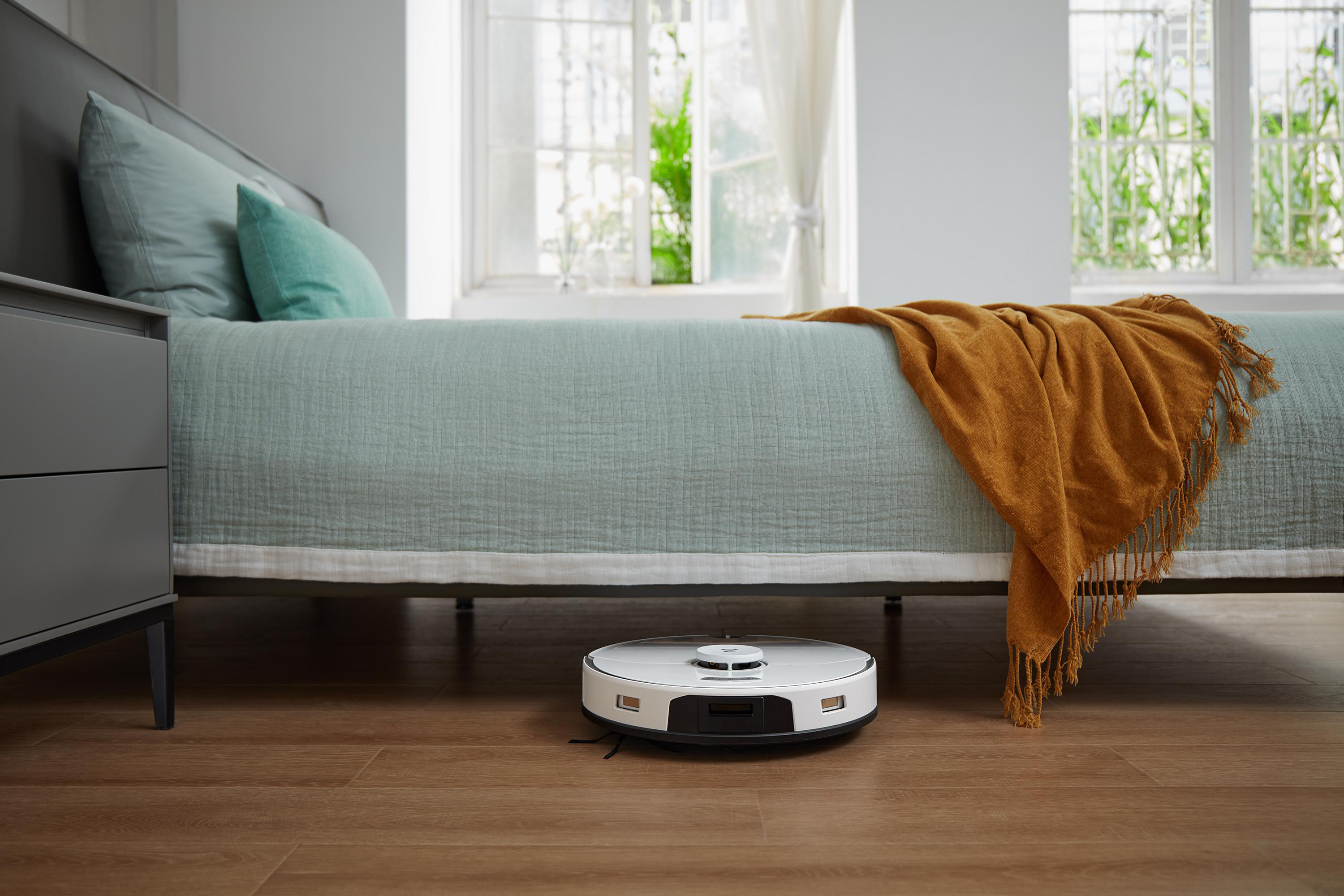 Ein weiß-schwarzer Roboter-Staubsauger auf einem Holzboden. Er steht in der Nähe eines Bettes mit einer Decke.