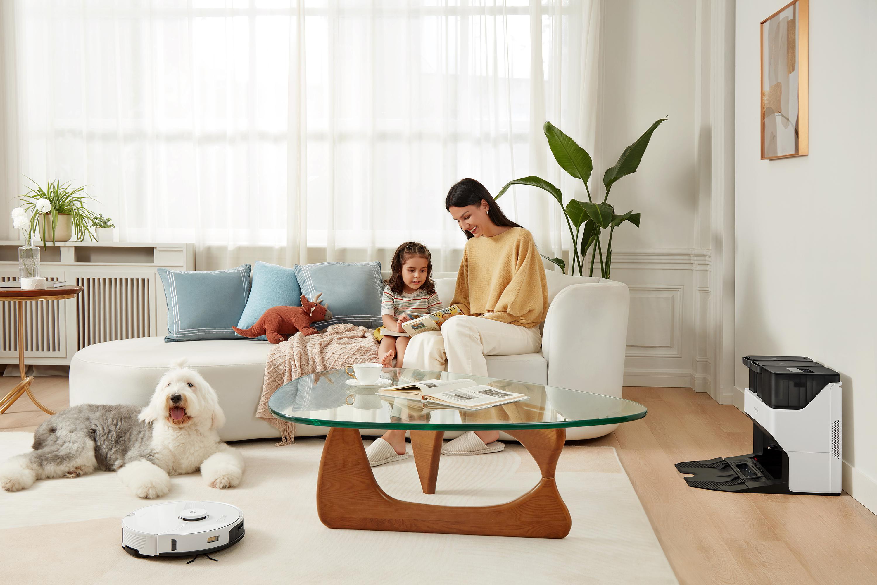 Eine Frau und ein Kind lesen auf einem Sofa, ein Hund und ein Roboterstaubsauger sind im Raum.