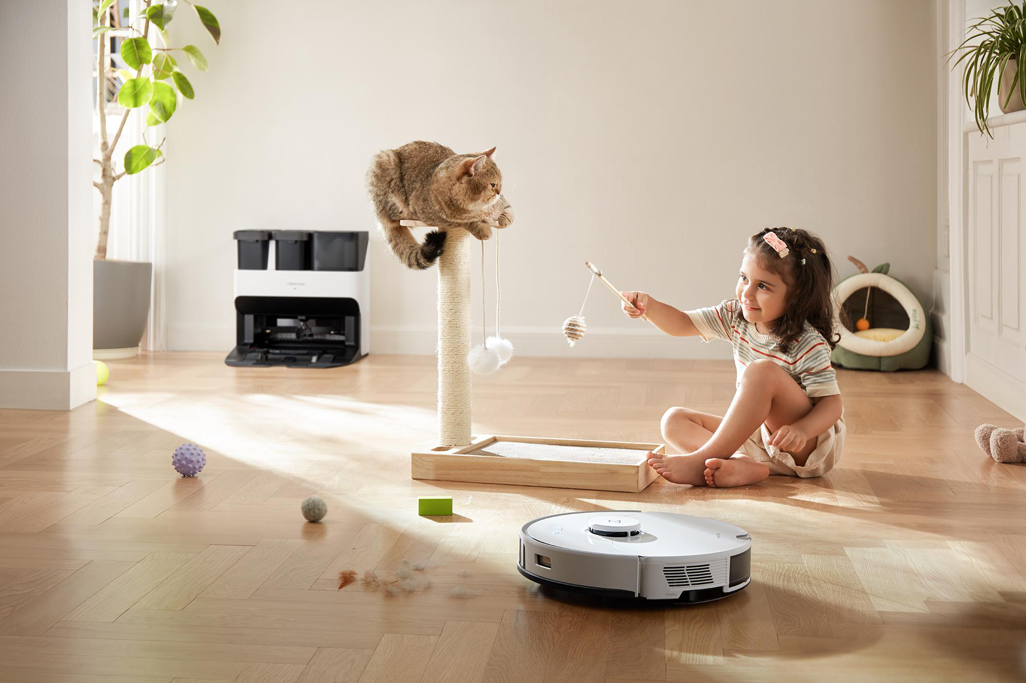 Ein Kind und eine Katze mit einem Kratzbaum und einem Roboter-Staubsauger. Die Katze ist auf dem Baum.