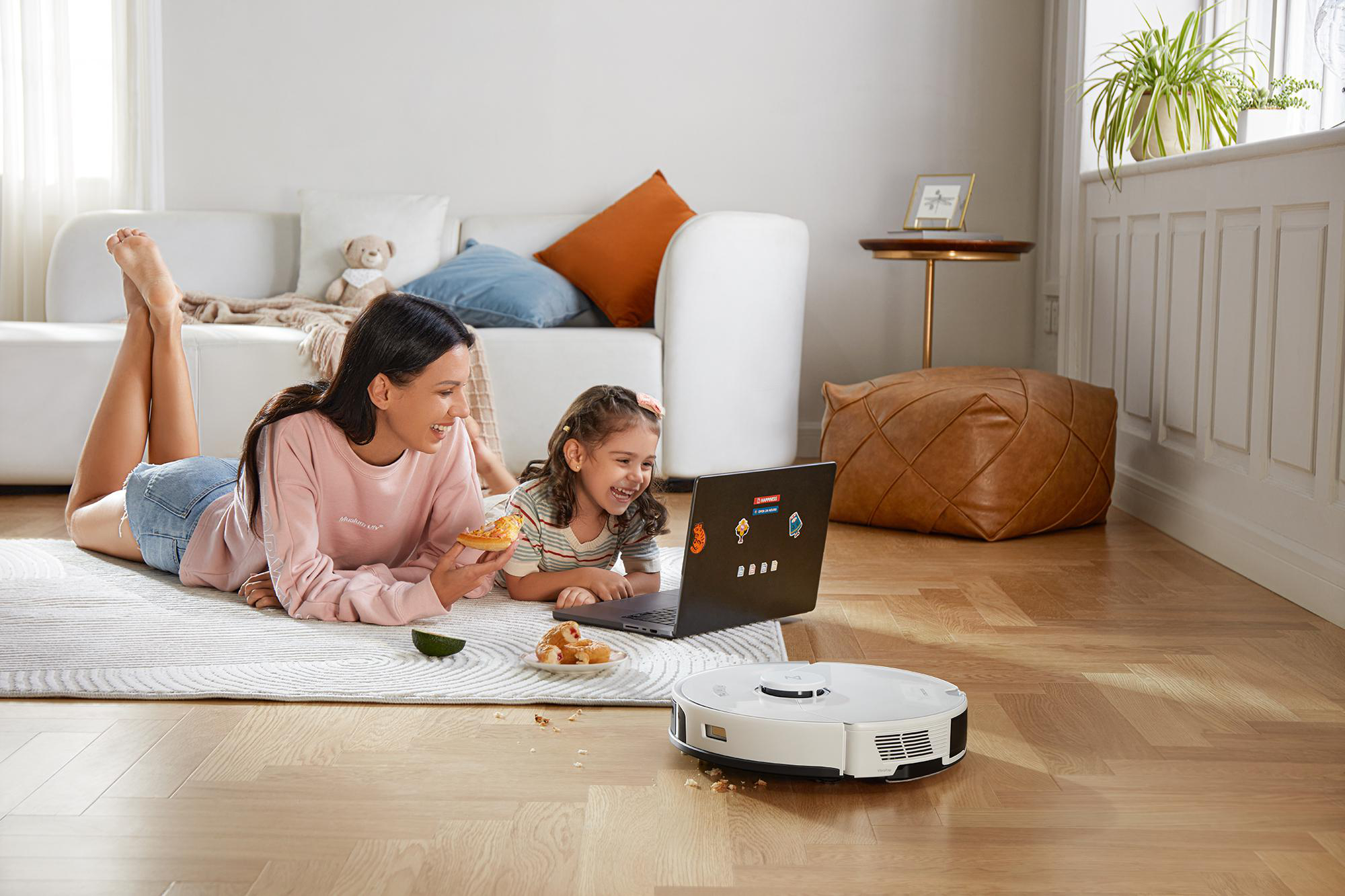 Mutter und Tochter sitzen auf einem Teppich. Ein Roboterstaubsauger steht auf dem Boden. Sie schauen auf einen Laptop.