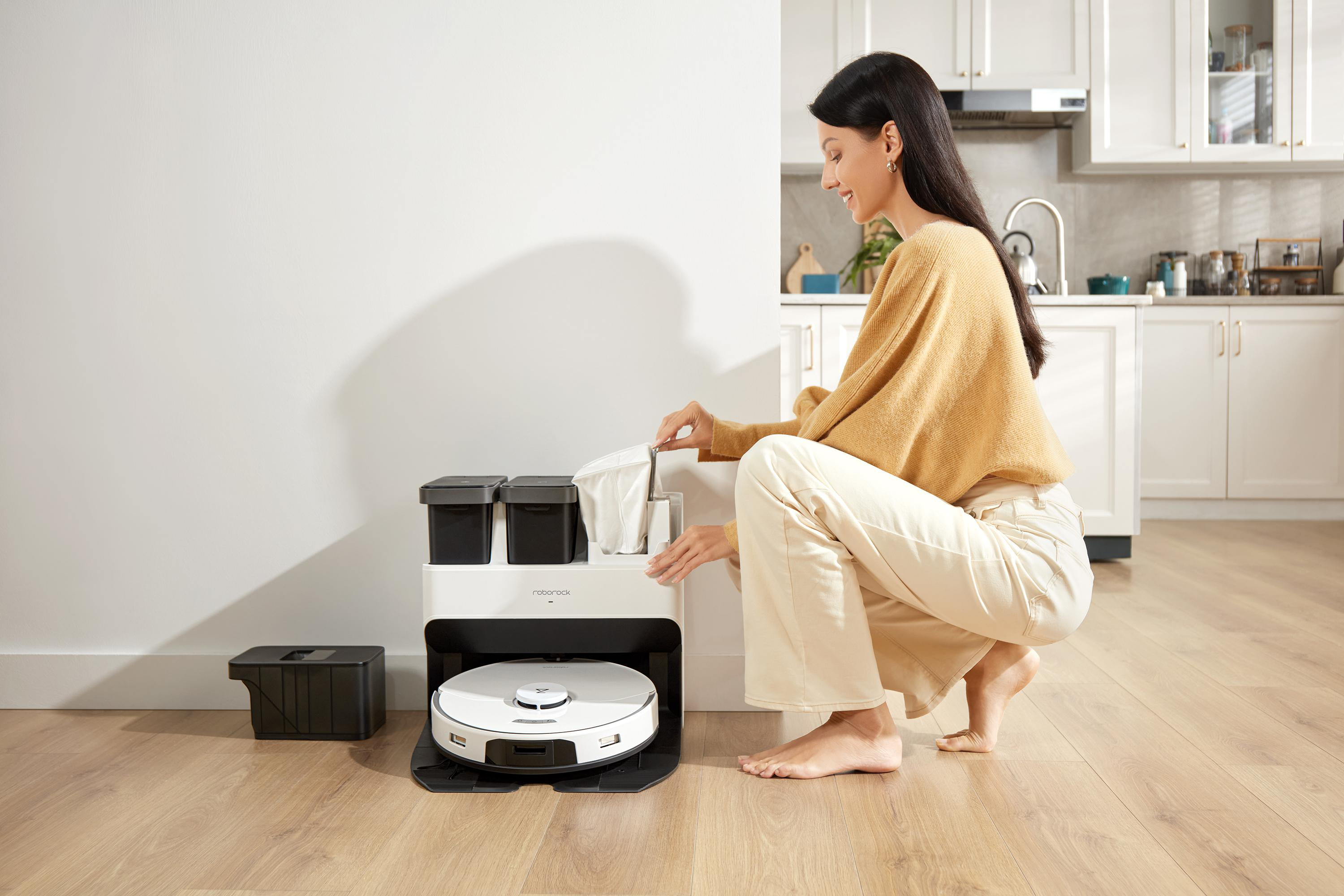 Eine Frau benutzt einen Roboter-Staubsauger in einer Küche. Der Staubsauger ist angedockt.