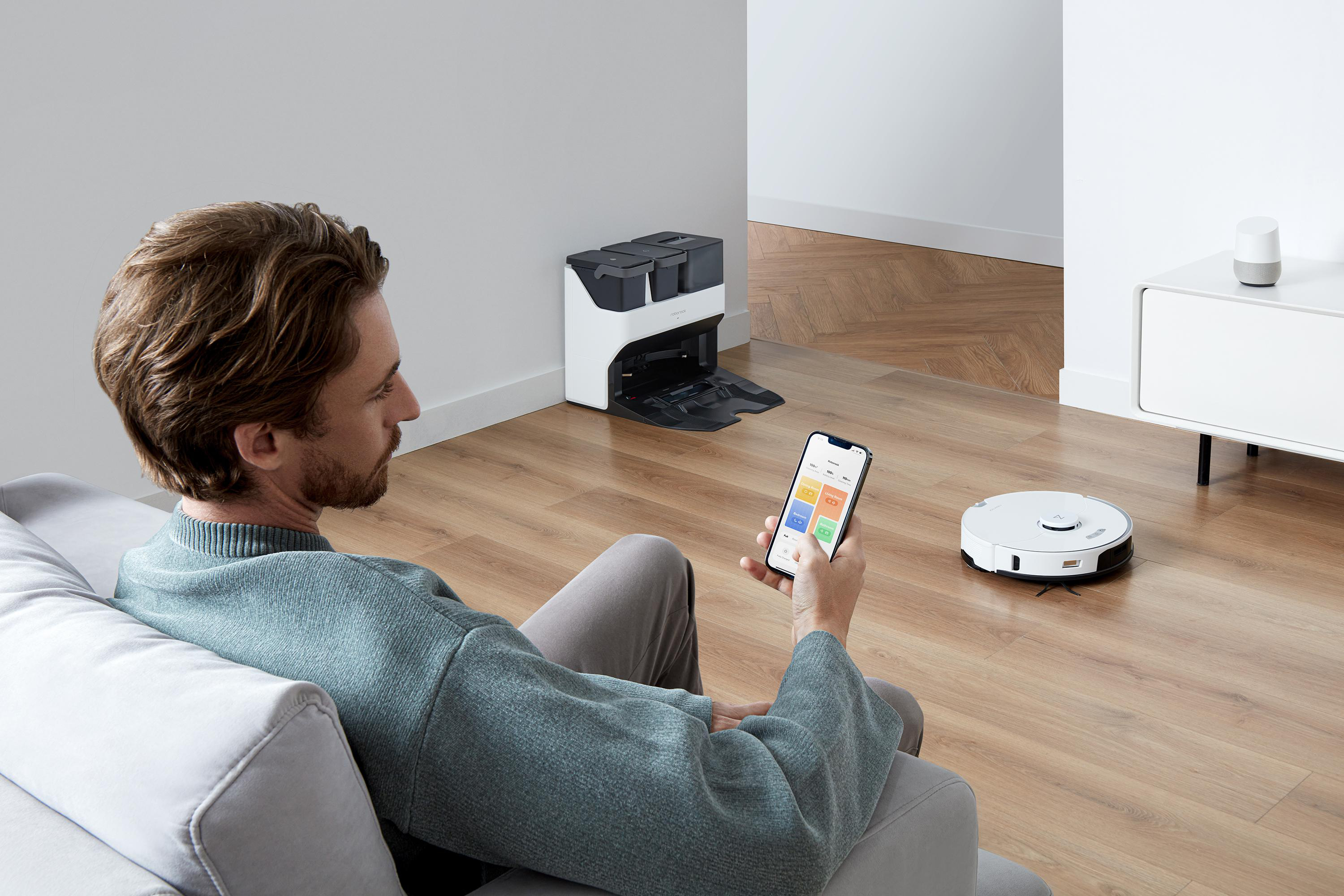 Mann mit Smartphone steuert Roboter-Staubsauger und Ladestation. Holzboden.