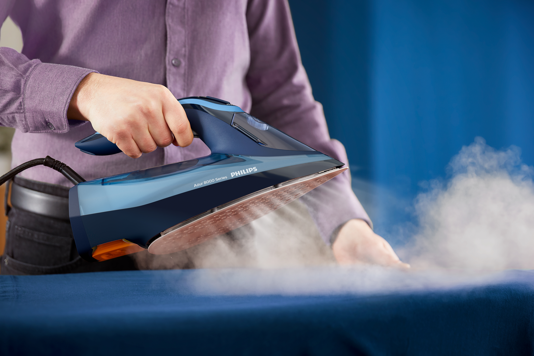 Person ironing blue fabric with a Philips iron. Steam is rising from the iron. Blue and gray iron.
