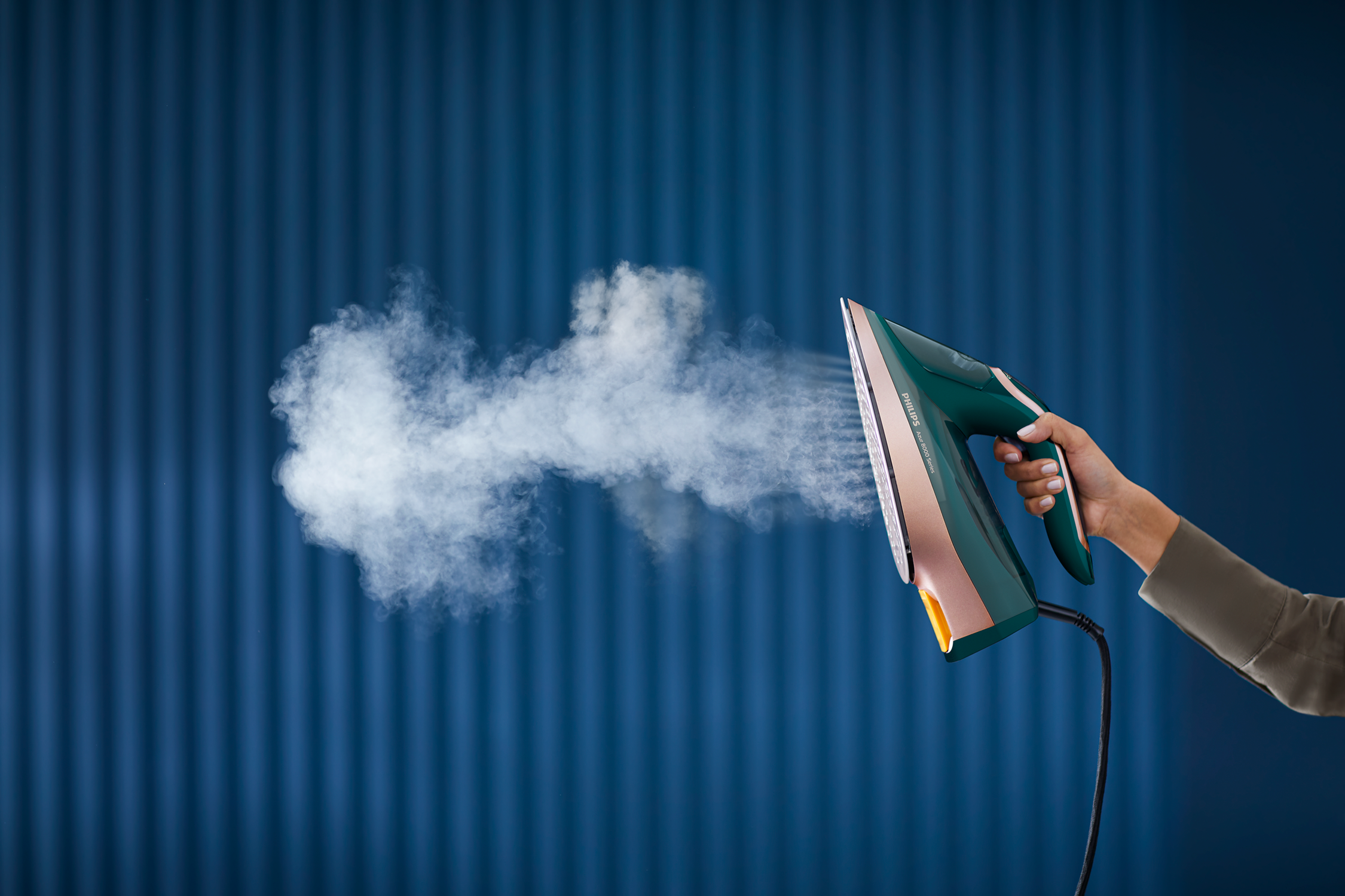 A person sprays steam from a green iron. The background is a blue wall.