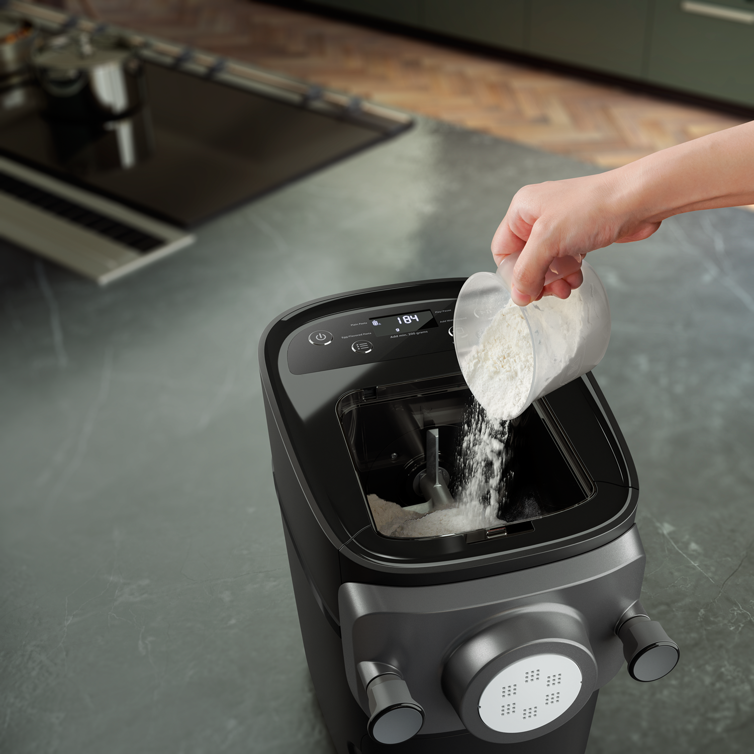 A hand pouring flour from a clear cup into a black pasta maker. The background is a kitchen countertop.