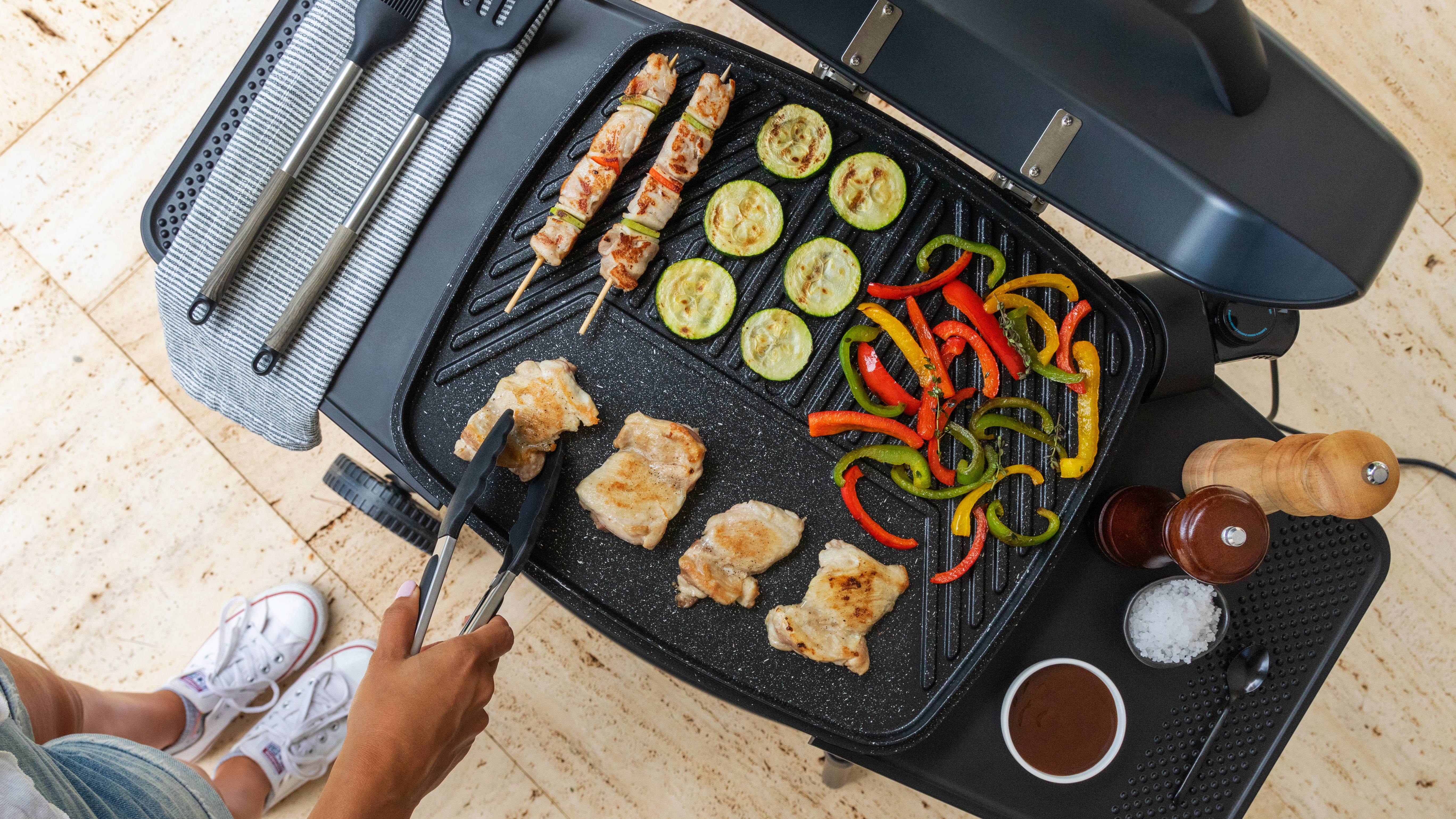 Parrilla con pollo, brochetas, verduras y pinzas. Una persona está asando. Ingredientes: calabacín, pimientos y salsa.