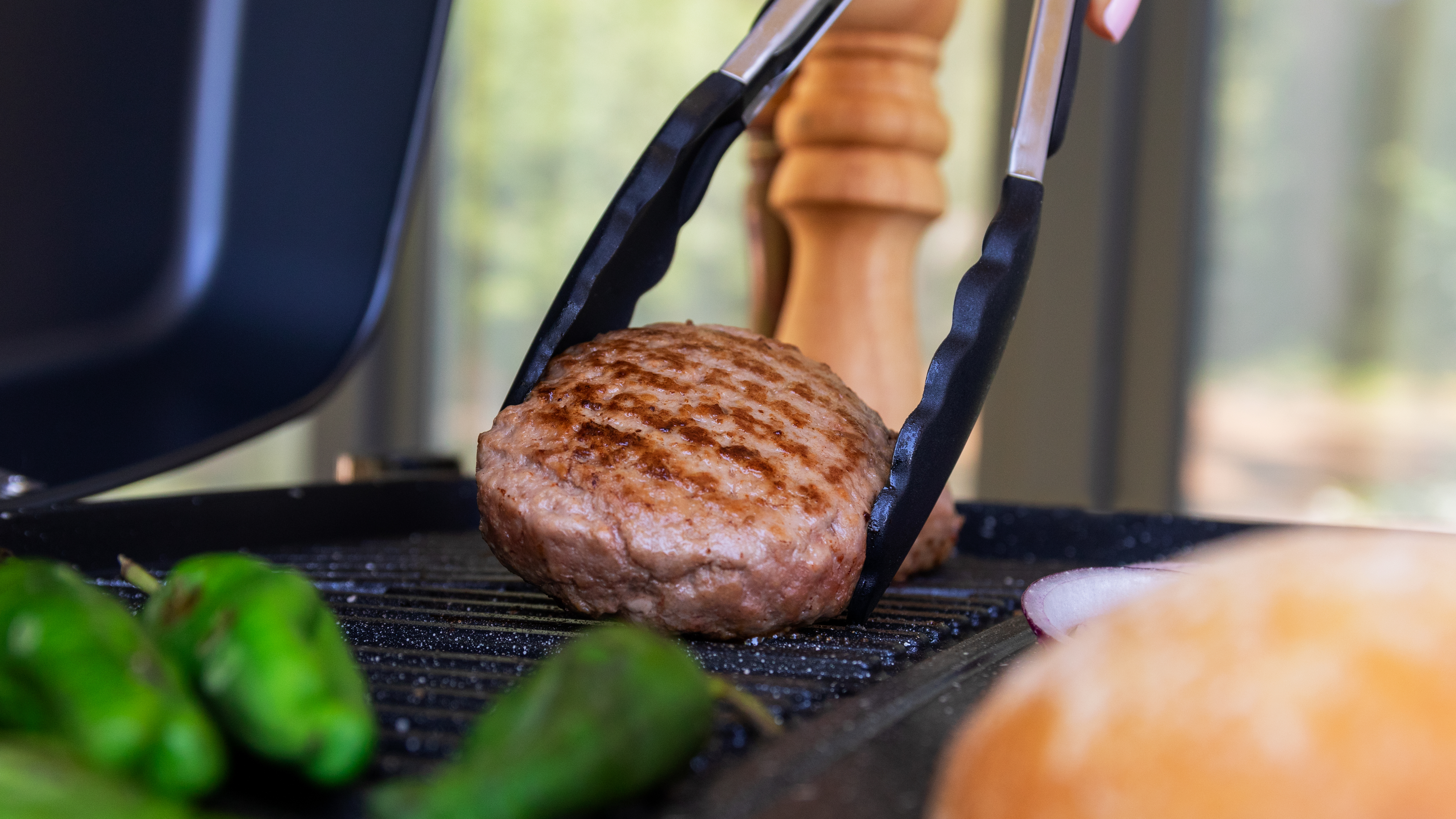 Una hamburguesa siendo levantada de la parrilla con pinzas, junto a pimientos y cebollas.