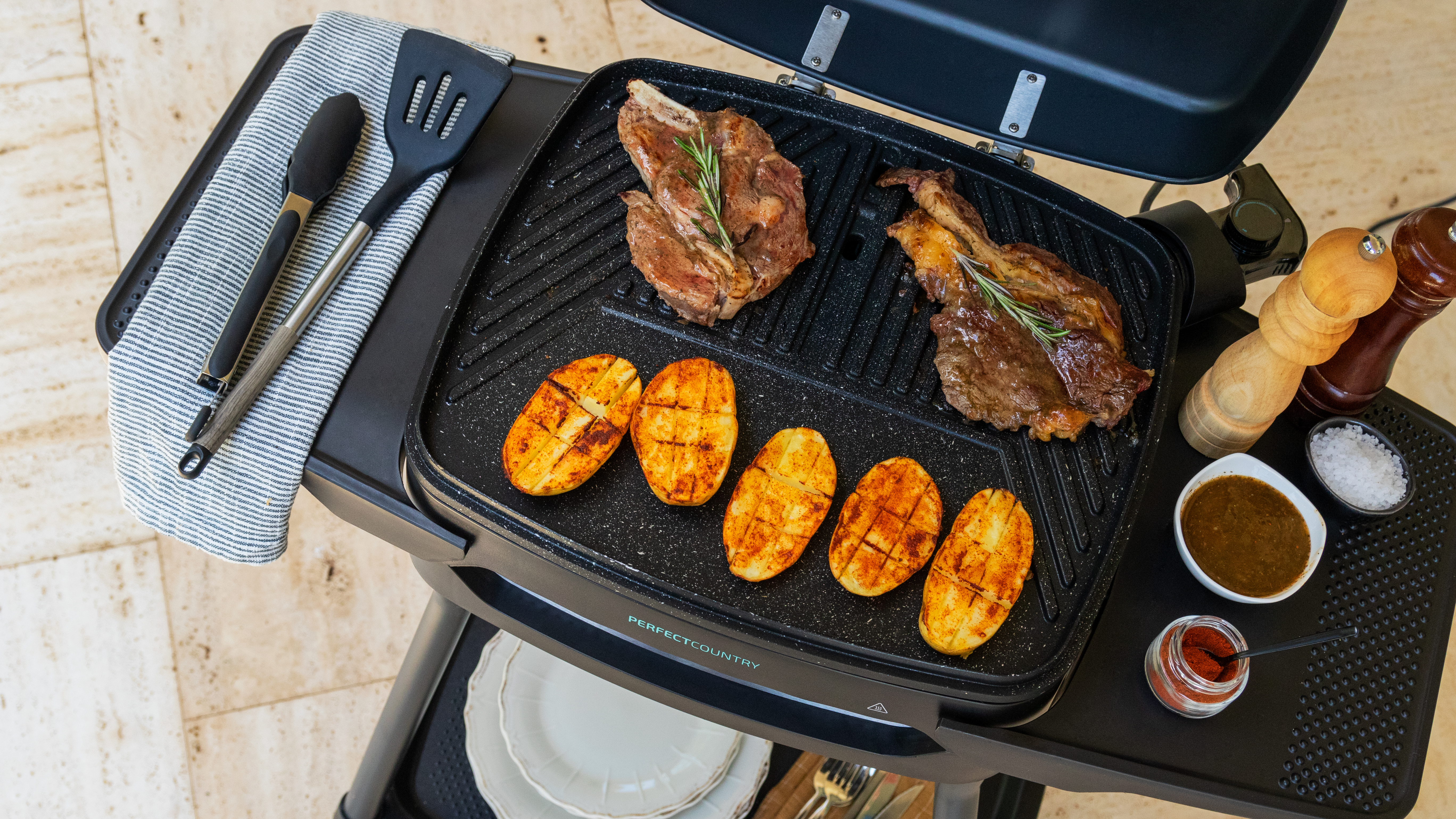 Parrilla con filetes y patatas. Pinzas y espátula a un lado. También hay sal, pimienta y salsas.
