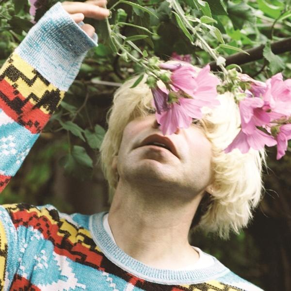 Homme blond avec des fleurs roses. Il porte un pull coloré. Les fleurs sont tenues près de son visage.