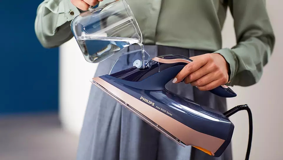 Une personne verse de l'eau d'une carafe dans un fer à repasser bleu et doré. Jupe grise et chemise verte.