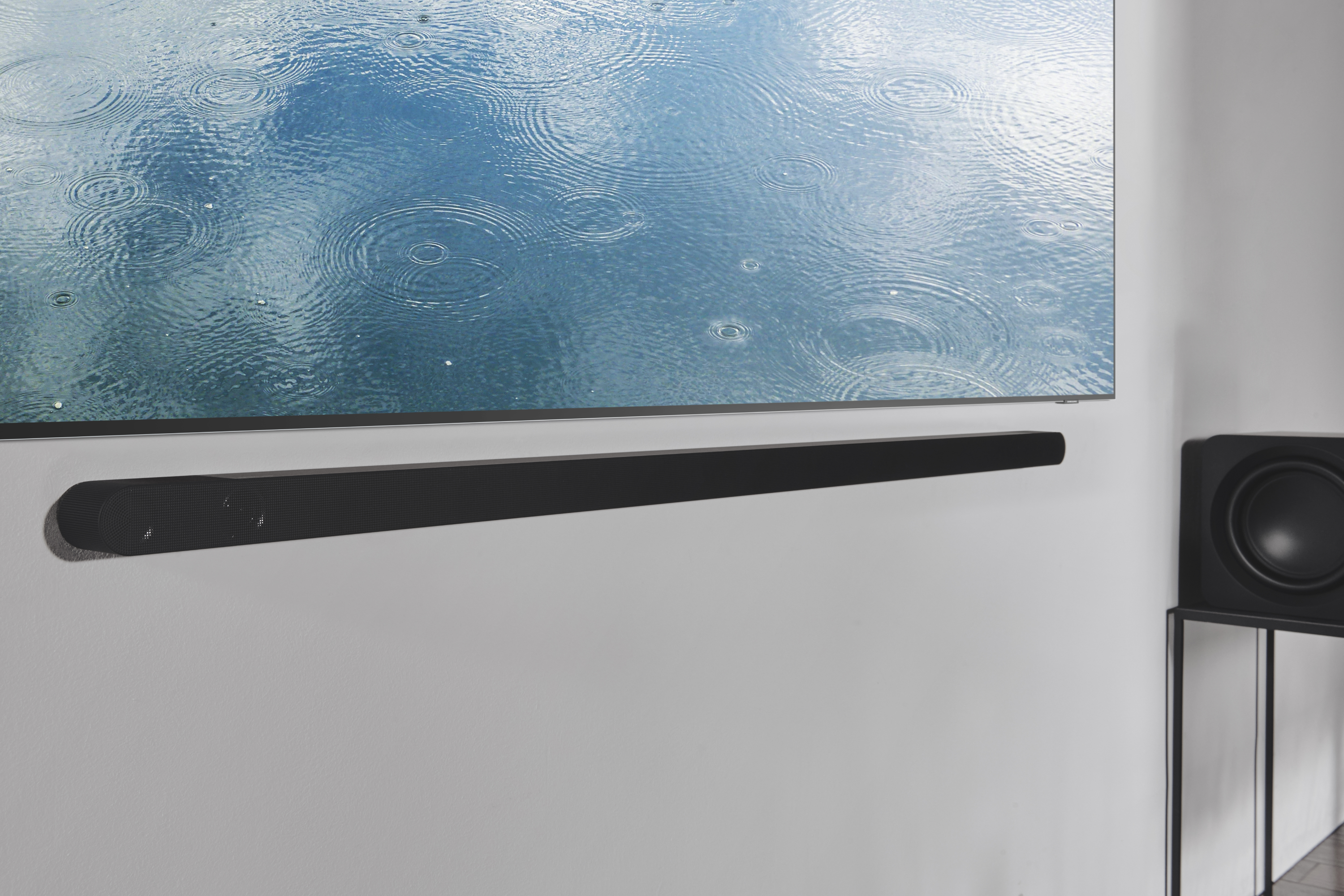 Una barra de sonido negra montada debajo de un televisor grande, en una pared blanca. Un subwoofer a la derecha.