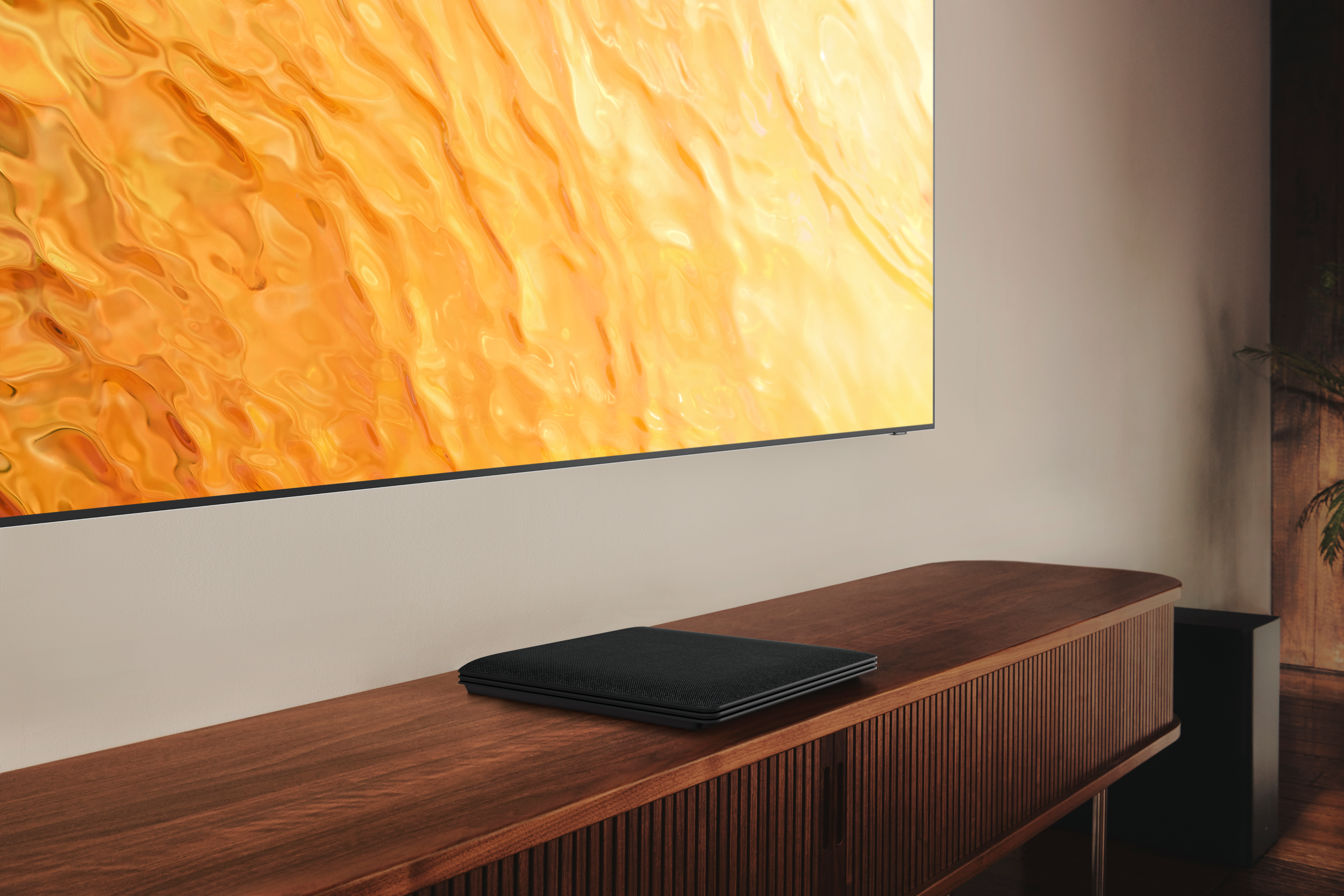 Un televisor está montado en una pared. Un dispositivo negro está colocado en un mueble de madera debajo del televisor.