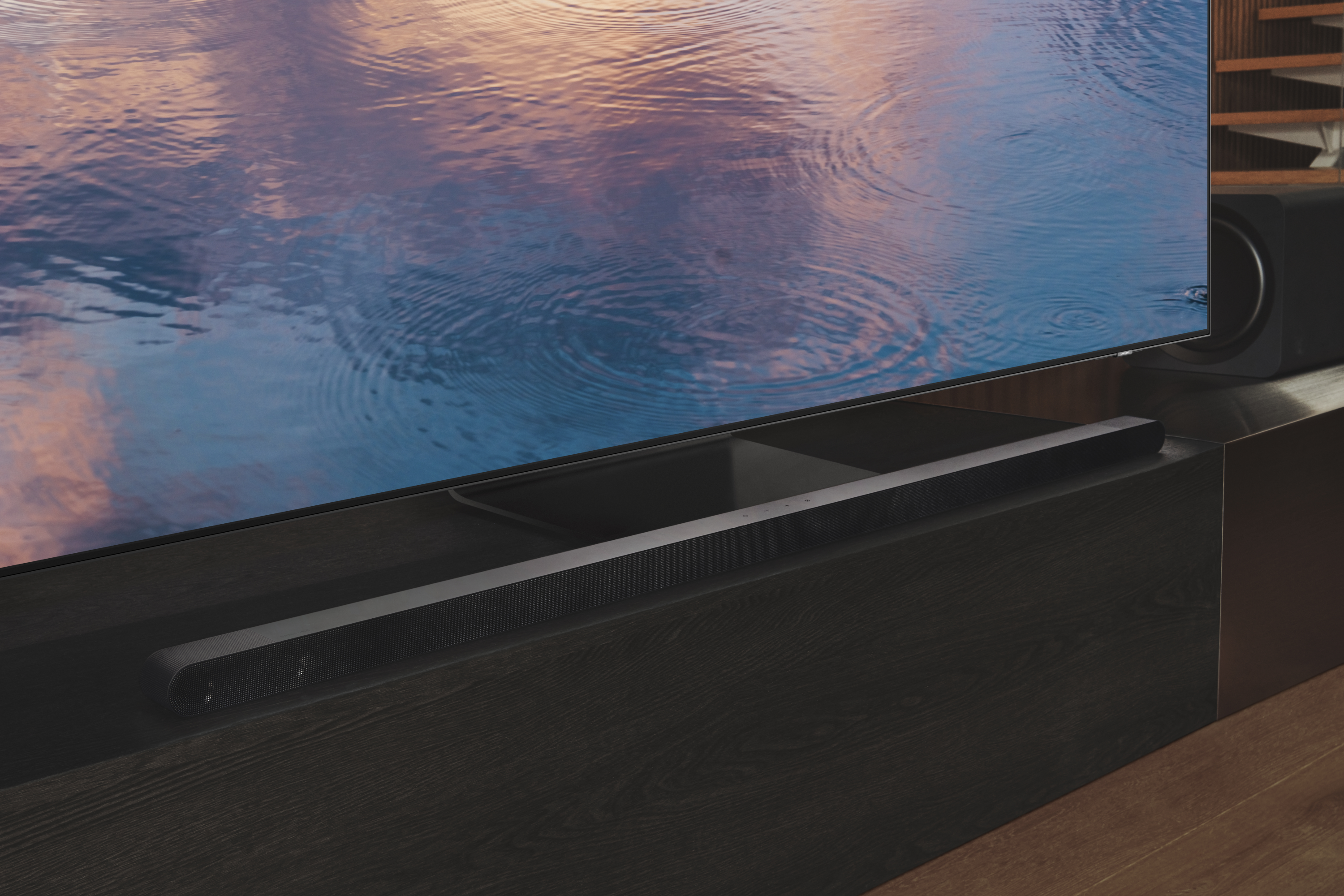 Barra de sonido negra sobre soporte de madera oscura debajo de un televisor grande. Reflejo de agua en la pantalla.