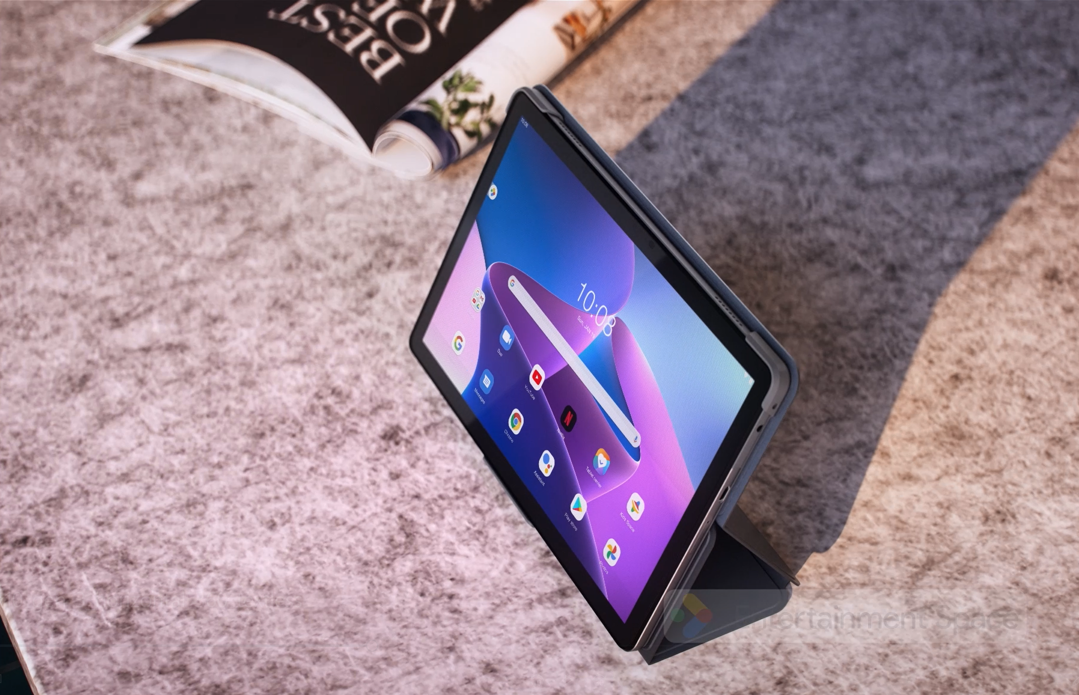 Tablet mit Uhr auf grauem Untergrund, daneben eine offene Zeitschrift.