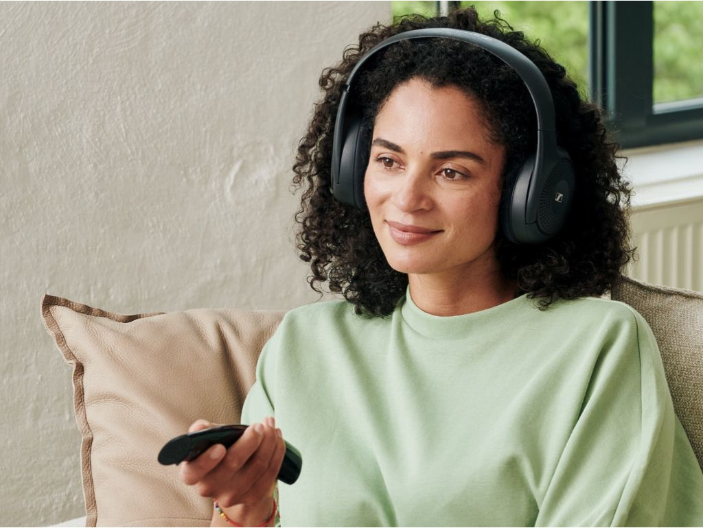 Woman wearing black headphones, sitting on a sofa, holding a remote, smiling.
