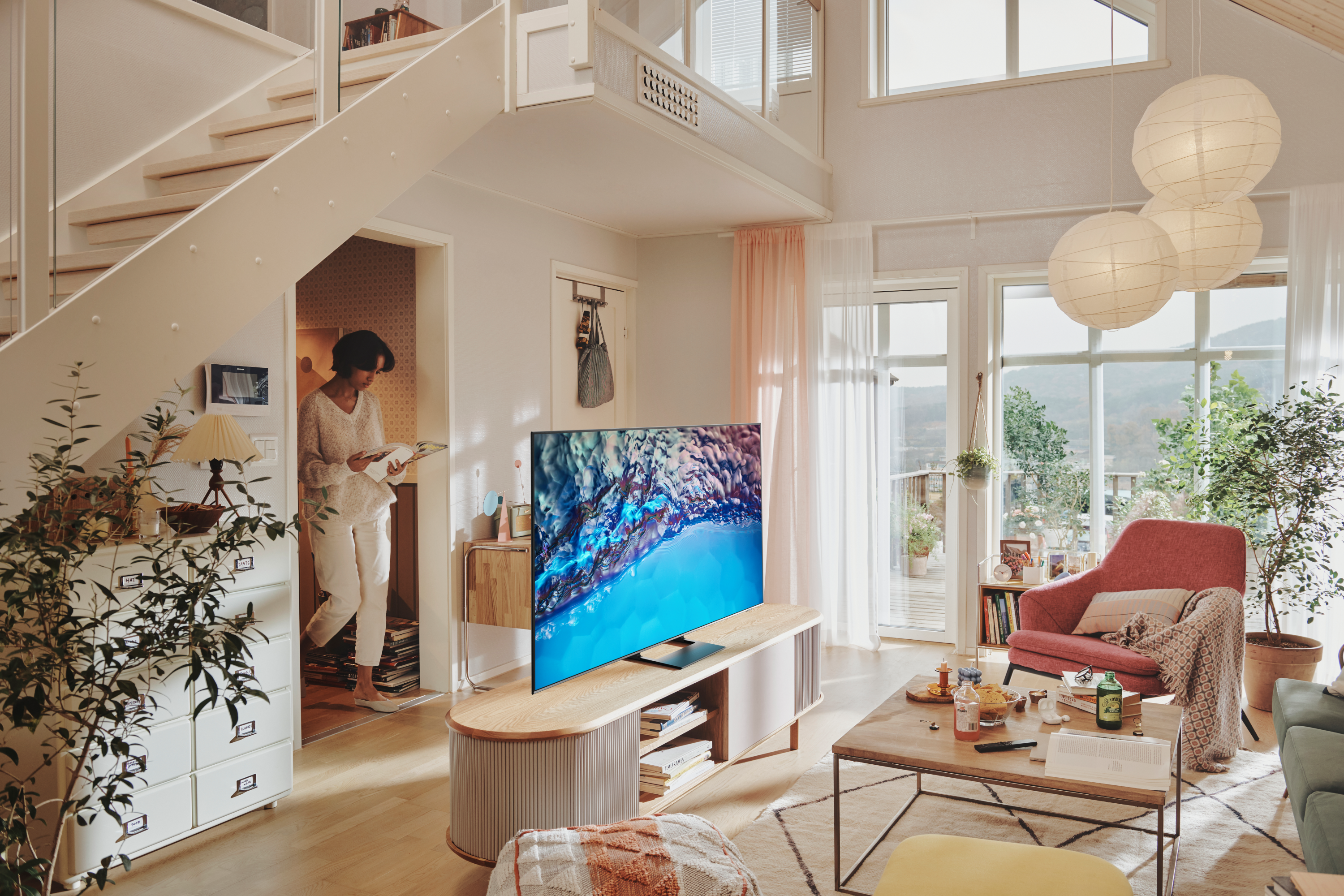 Una mujer en una sala luminosa. Un gran televisor sobre un soporte. Escaleras y ventanas visibles.