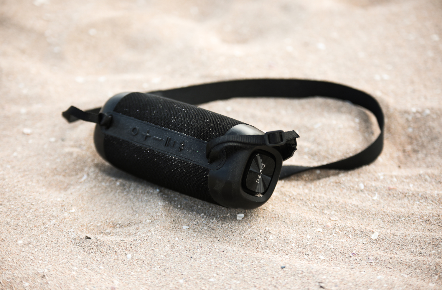A black portable speaker with a strap on a sandy beach. The speaker is cylindrical.
