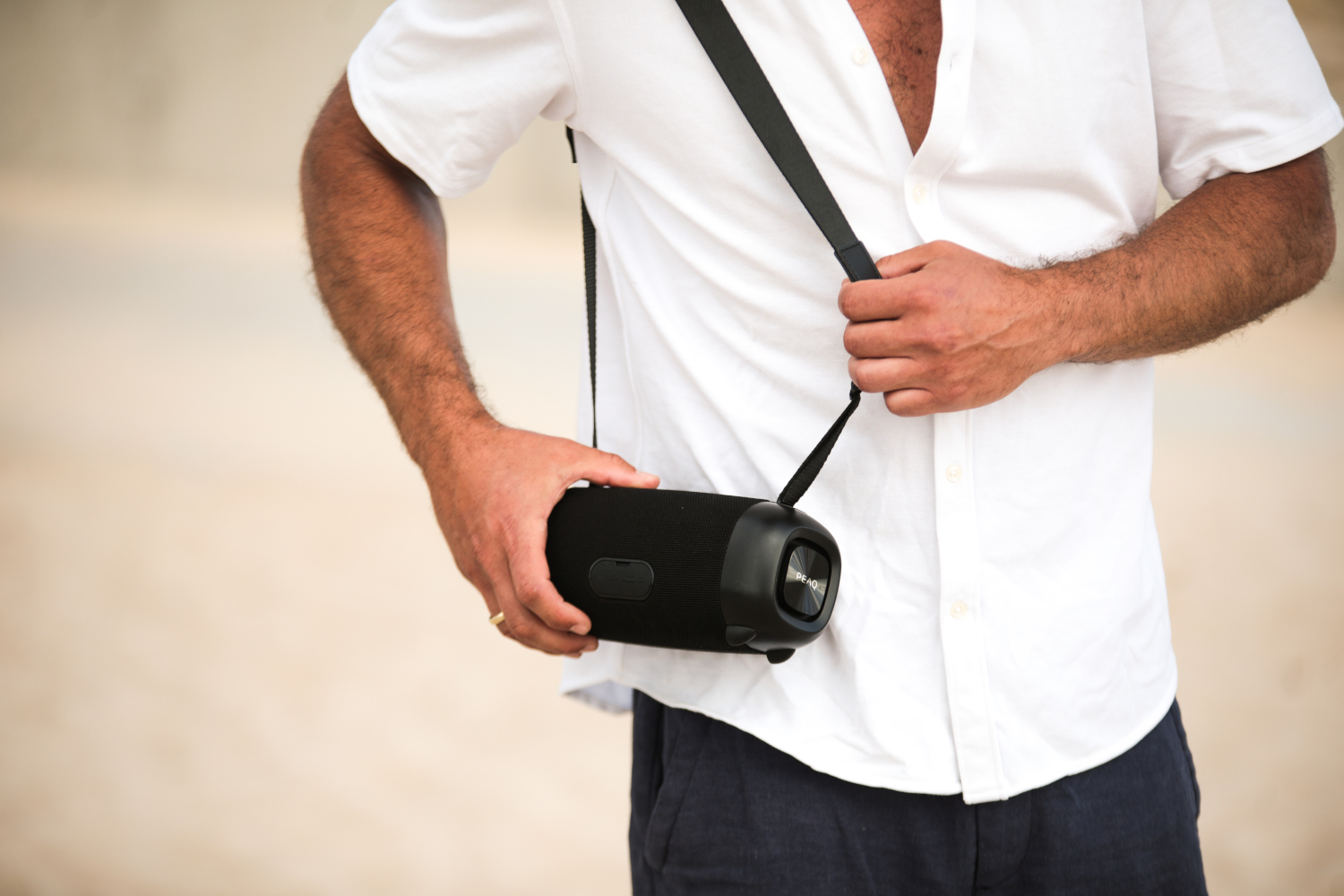 A man in a white shirt holds a black portable speaker with a strap over his shoulder.