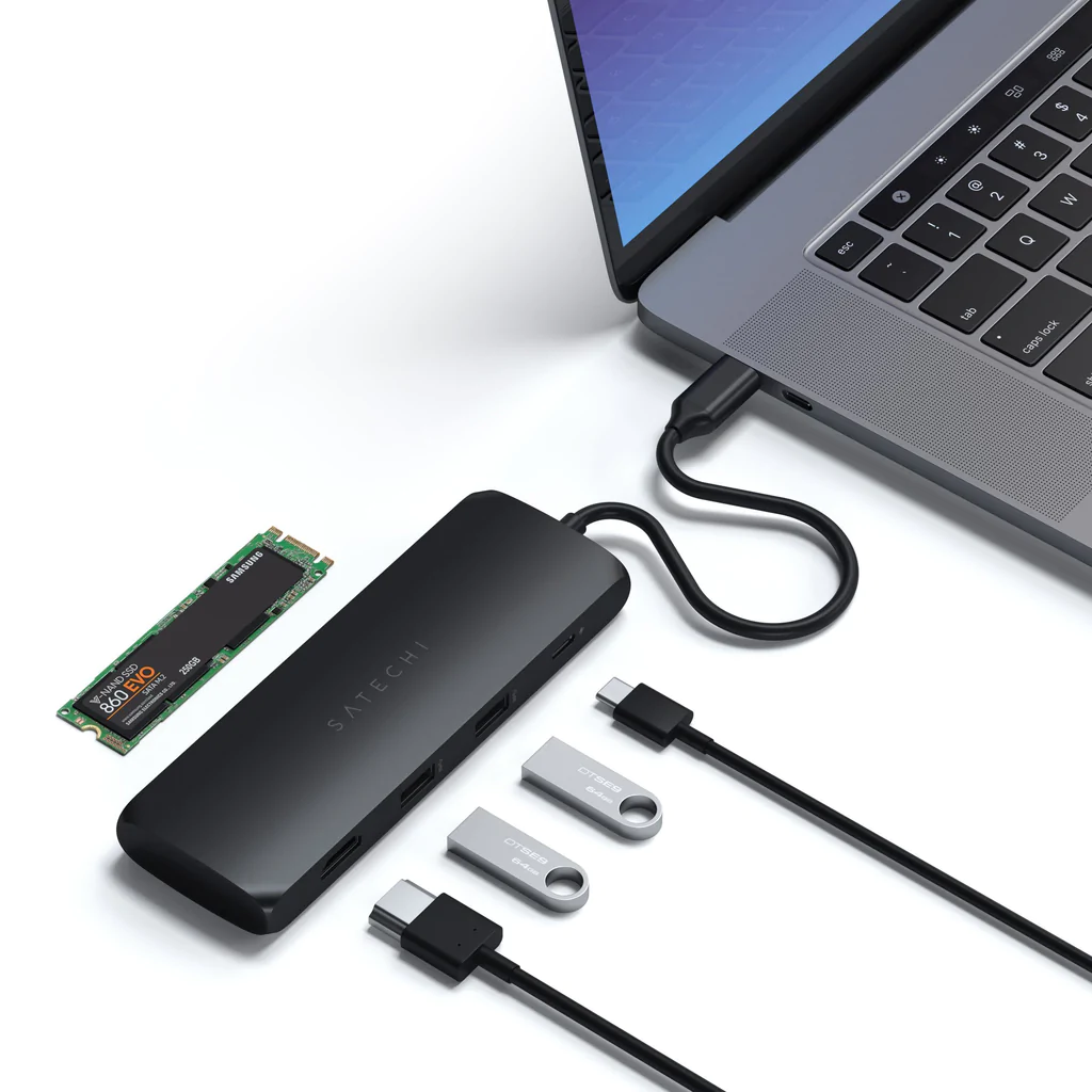 Egy laptop egy fekete Satechi hubhoz csatlakoztatva. USB meghajtók és egy SSD is látható.
