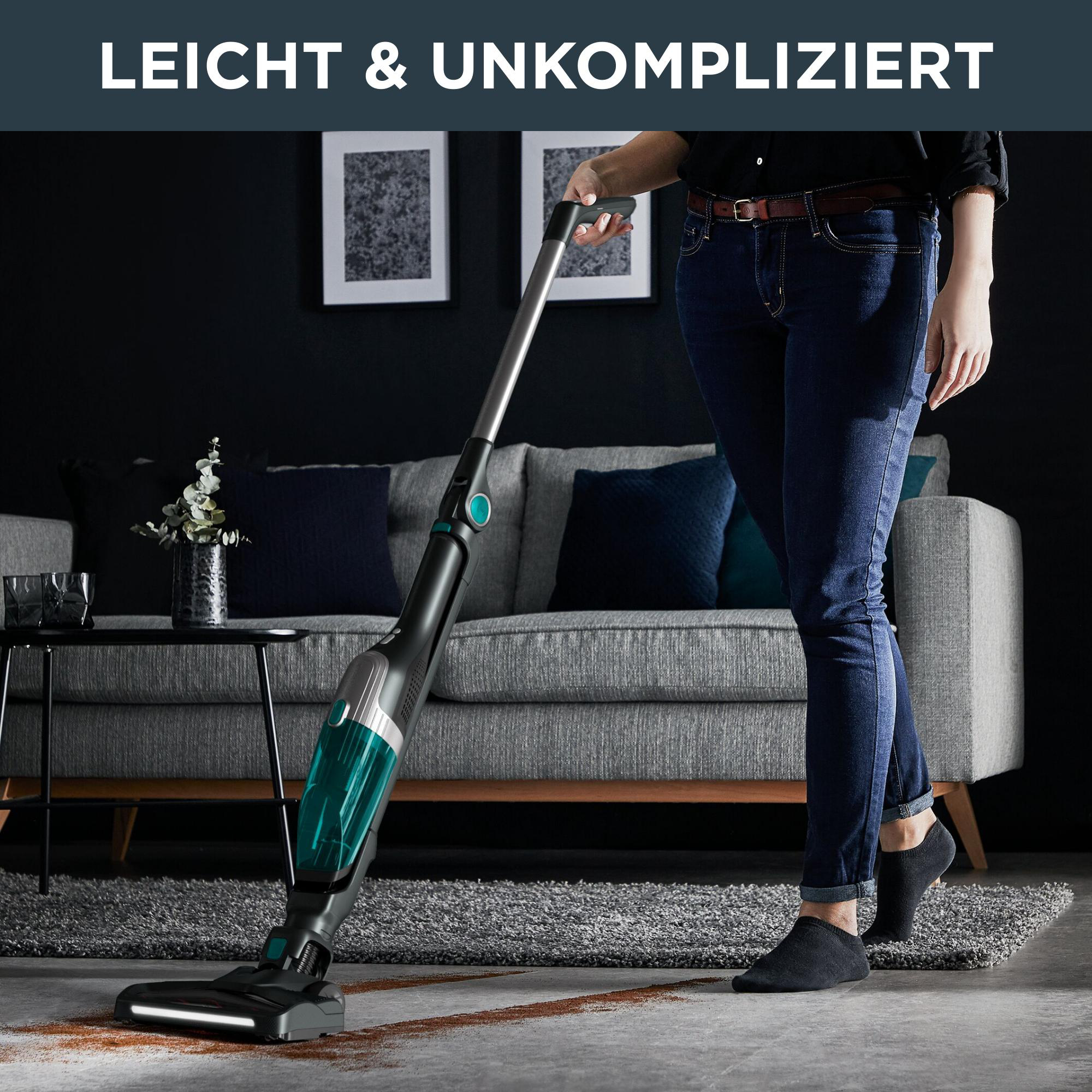 Eine Frau, die einen Raum staubsaugt, mit einem kabellosen Staubsauger auf einem Teppich in der Nähe eines grauen Sofas.
