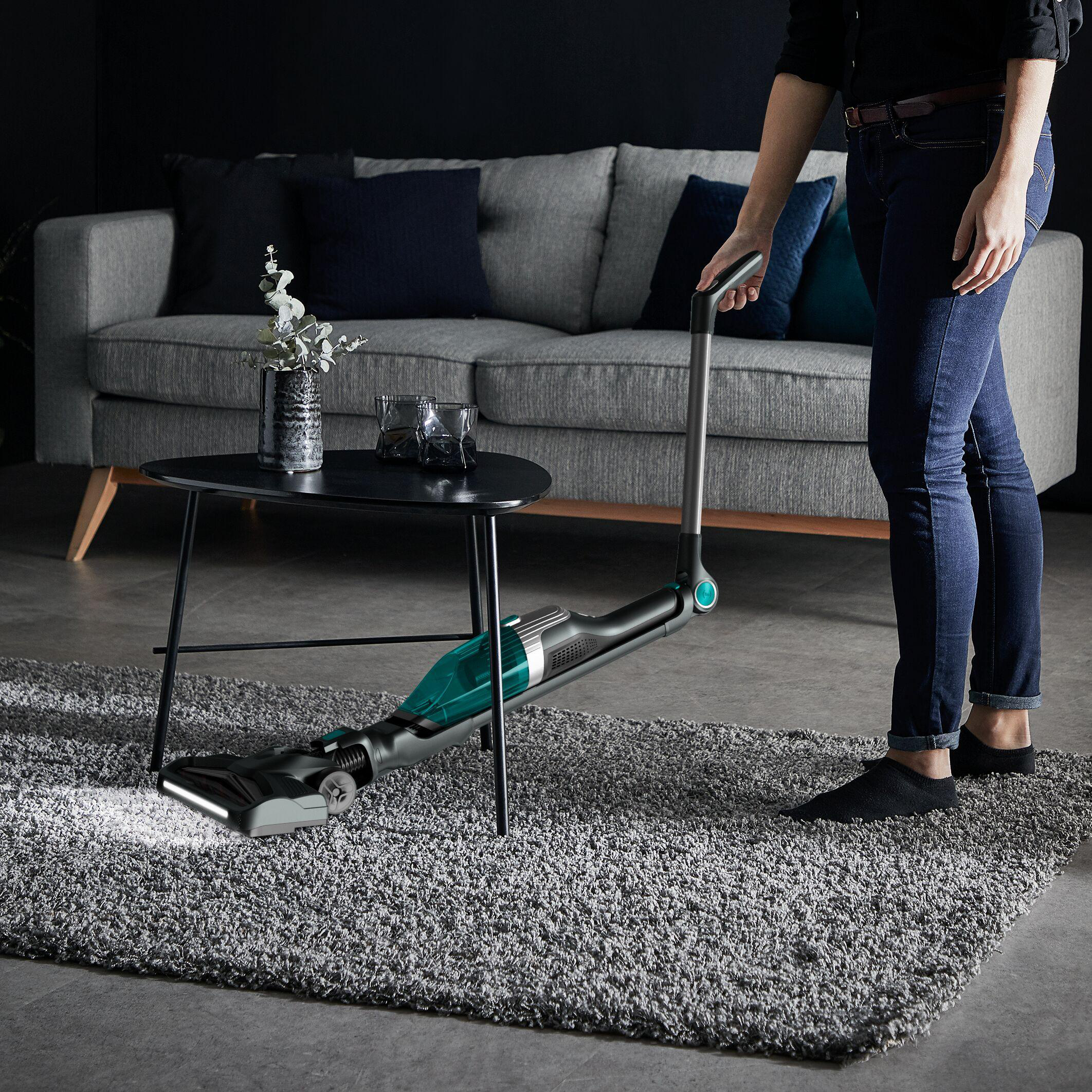 Person saugt grauen Teppich mit einem kabellosen Staubsauger. Schwarzer Tisch und Sofa im Hintergrund.