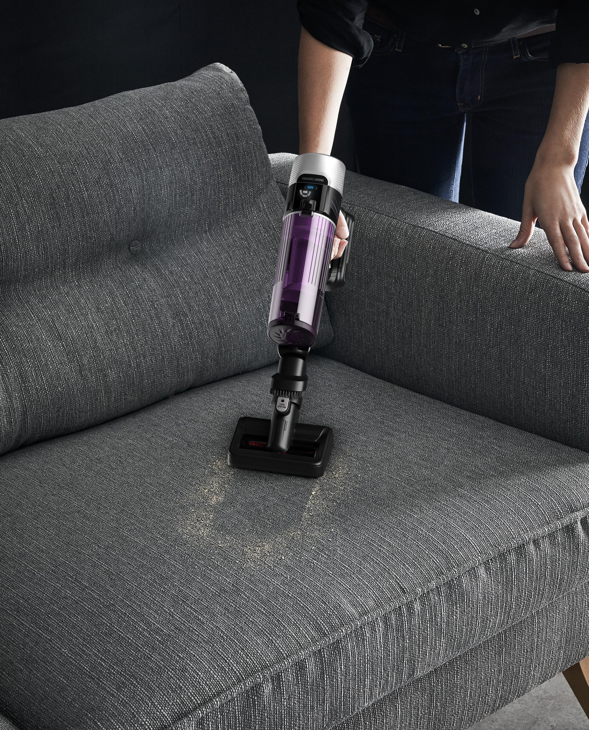Eine Person saugt ein graues Sofa mit einem Handstaubsauger. Staub wird aufgesaugt. Das Sofa hat Knöpfe.