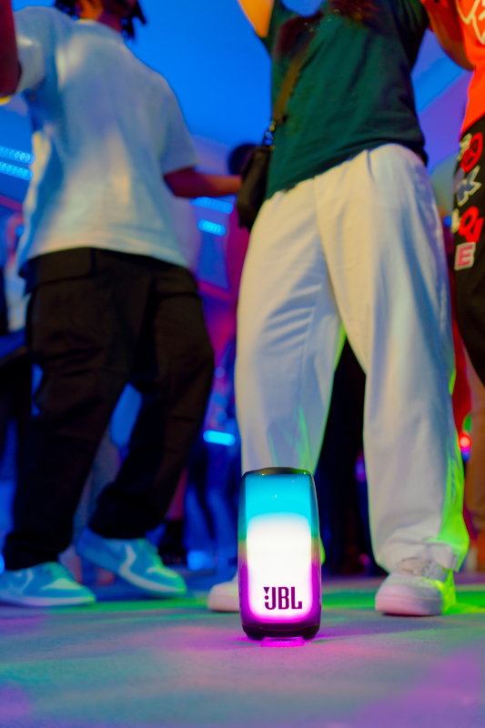 Eine Tanzszene mit einem JBL-Lautsprecher. Menschen tanzen mit bunten Lichtern im Hintergrund.