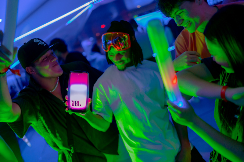 Gruppe von Leuten auf einer Party mit Neonlichtern. Eine Person hält einen JBL-Lautsprecher, andere haben Leuchtstäbe.