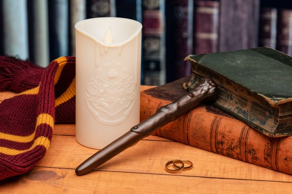 Kerze, Zauberstab, Bücher, Schal und Ringe auf einem Holztisch. Bücherregal im Hintergrund.