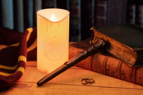 Brennende Kerze mit Hogwarts-Wappen, Zauberstab, Bücher und Schal auf Holztisch. Gemütliche Szene.